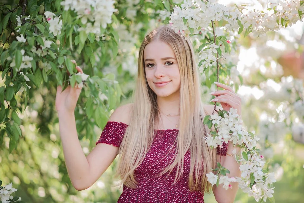 A young woman with long blonde hair standing among flowering trees with white blossoms, wearing an off-the-shoulder maroon dress.