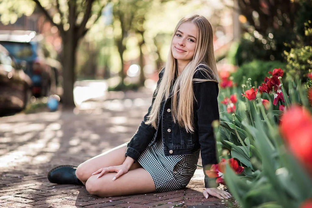A young woman with long blonde hair sitting on a brick sidewalk next to a flower bed with red flowers, smiling at the camera.