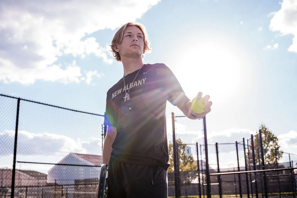 Young man with long hair playing tennis outdoors during daytime, holding a tennis ball, wearing a black shirt that says 'New Albany.'
