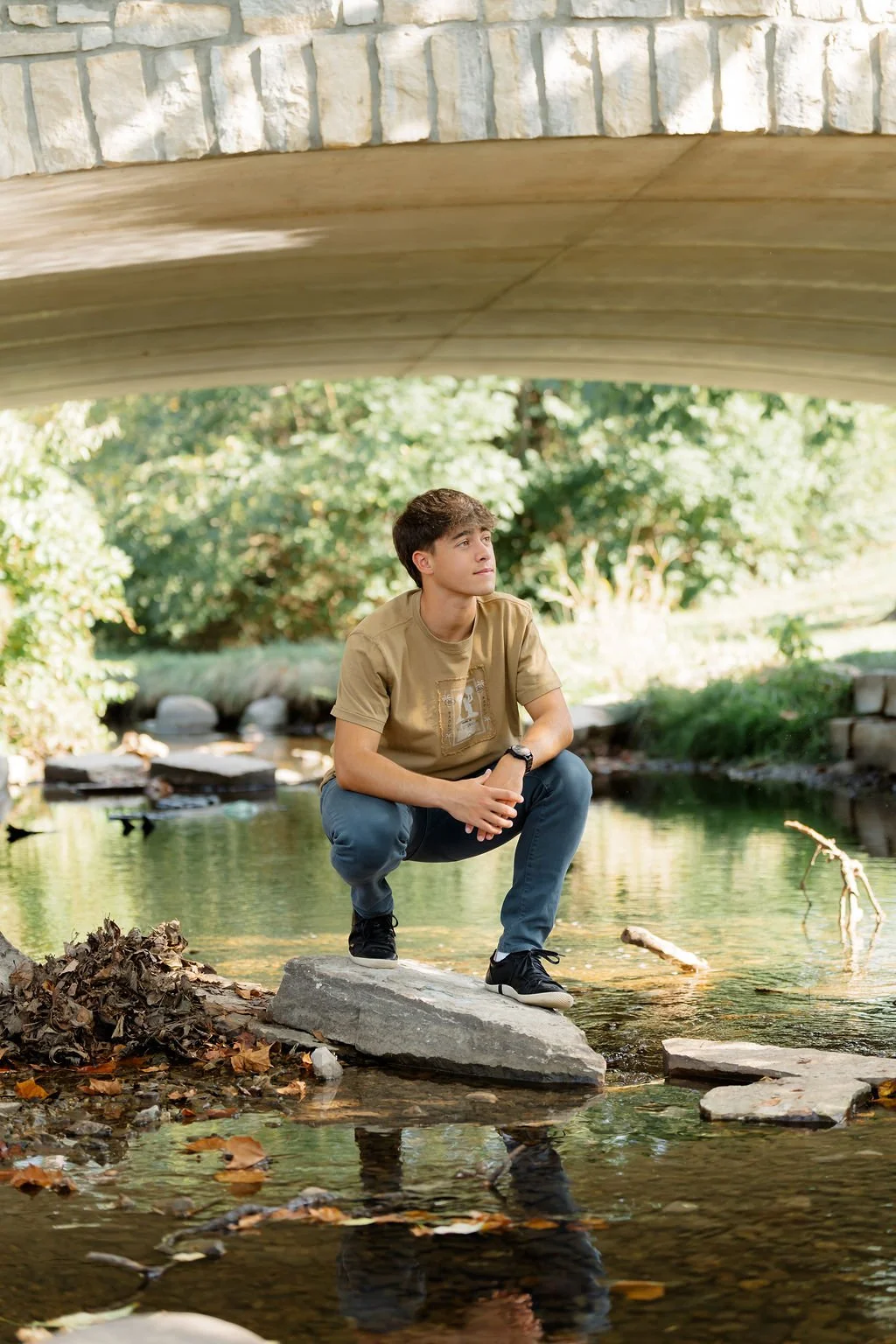 A young man with brown hair crouches on a rock in a creek, looking thoughtfully into the distance. The scene is outdoors with lush green trees and under a bridge.