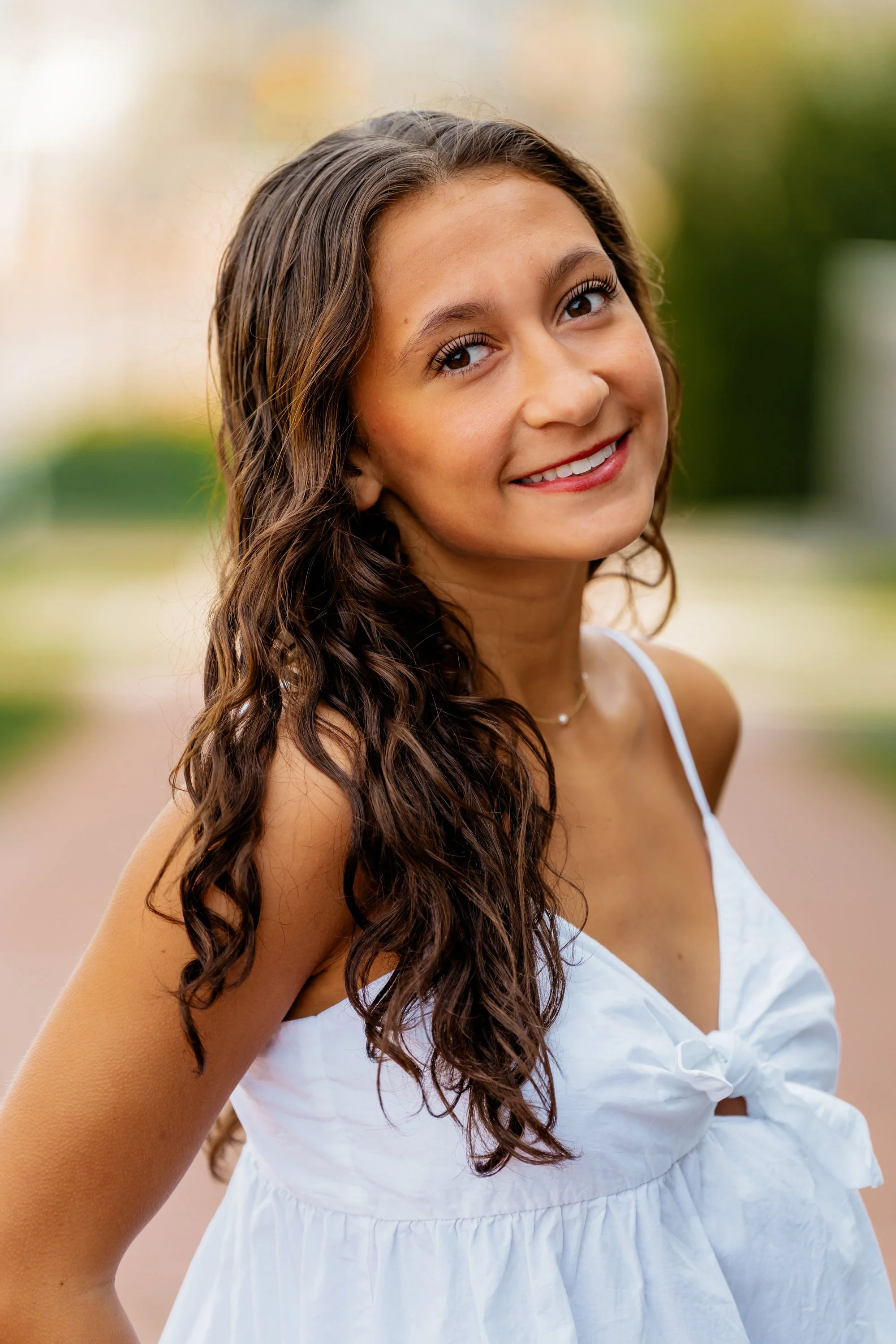A young woman with long, curly brown hair wearing a white dress and smiling outdoors.