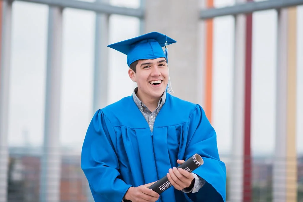 A young man in a blue graduation cap and gown holding a diploma, smiling in a modern building with large windows.