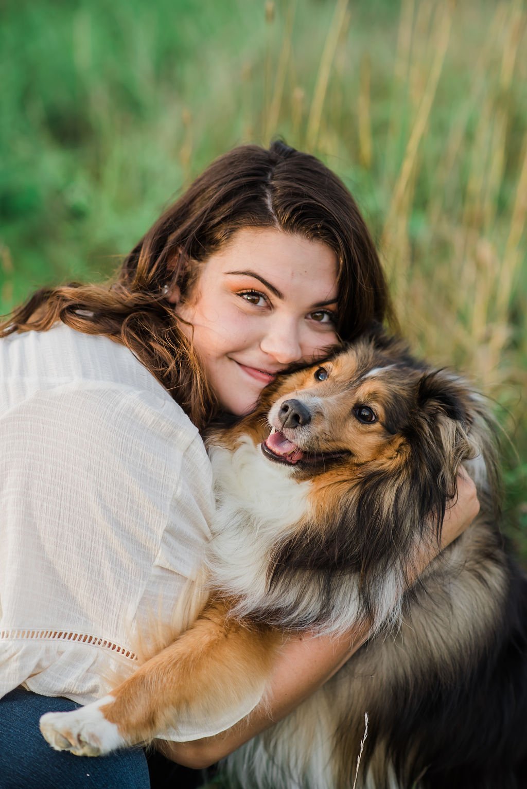 A woman hugging a fluffy Australian Shepherd dog outdoors with tall grass in the background.