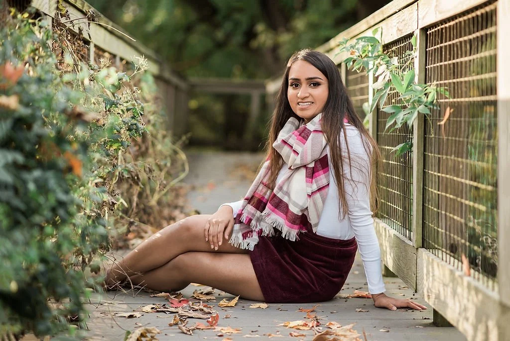 A young woman with long dark hair sitting on a wooden pathway outdoors, surrounded by autumn leaves and greenery, wearing a white top, maroon skirt, and a plaid scarf.