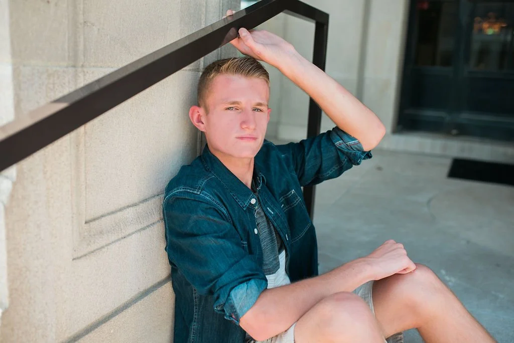 Young man sitting against a beige wall outside, looking at the camera with a serious expression, wearing a denim shirt and shorts.