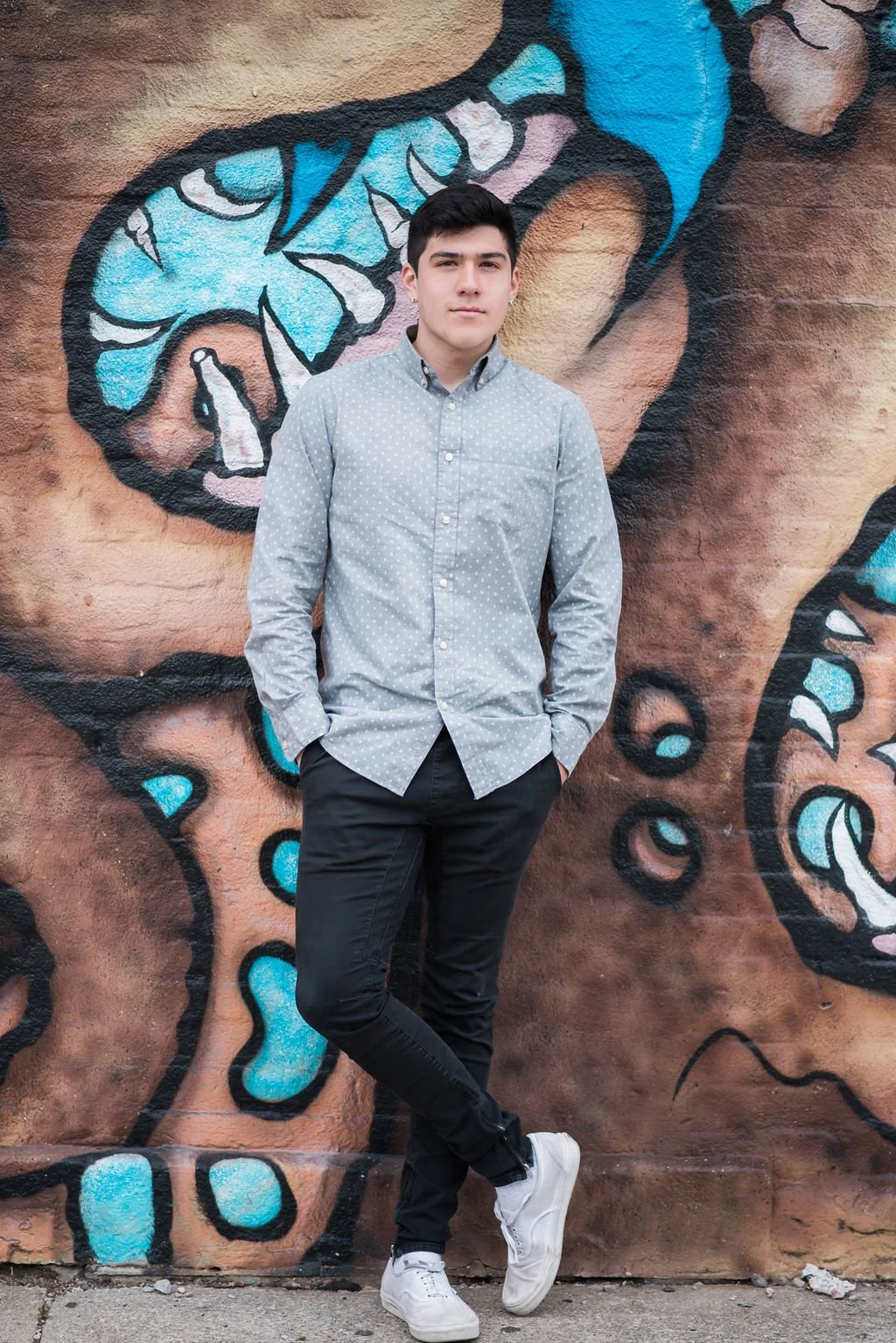 A young man standing casually with hands in pockets in front of a colorful mural on a brick wall, featuring abstract designs in blue, white, and pink.