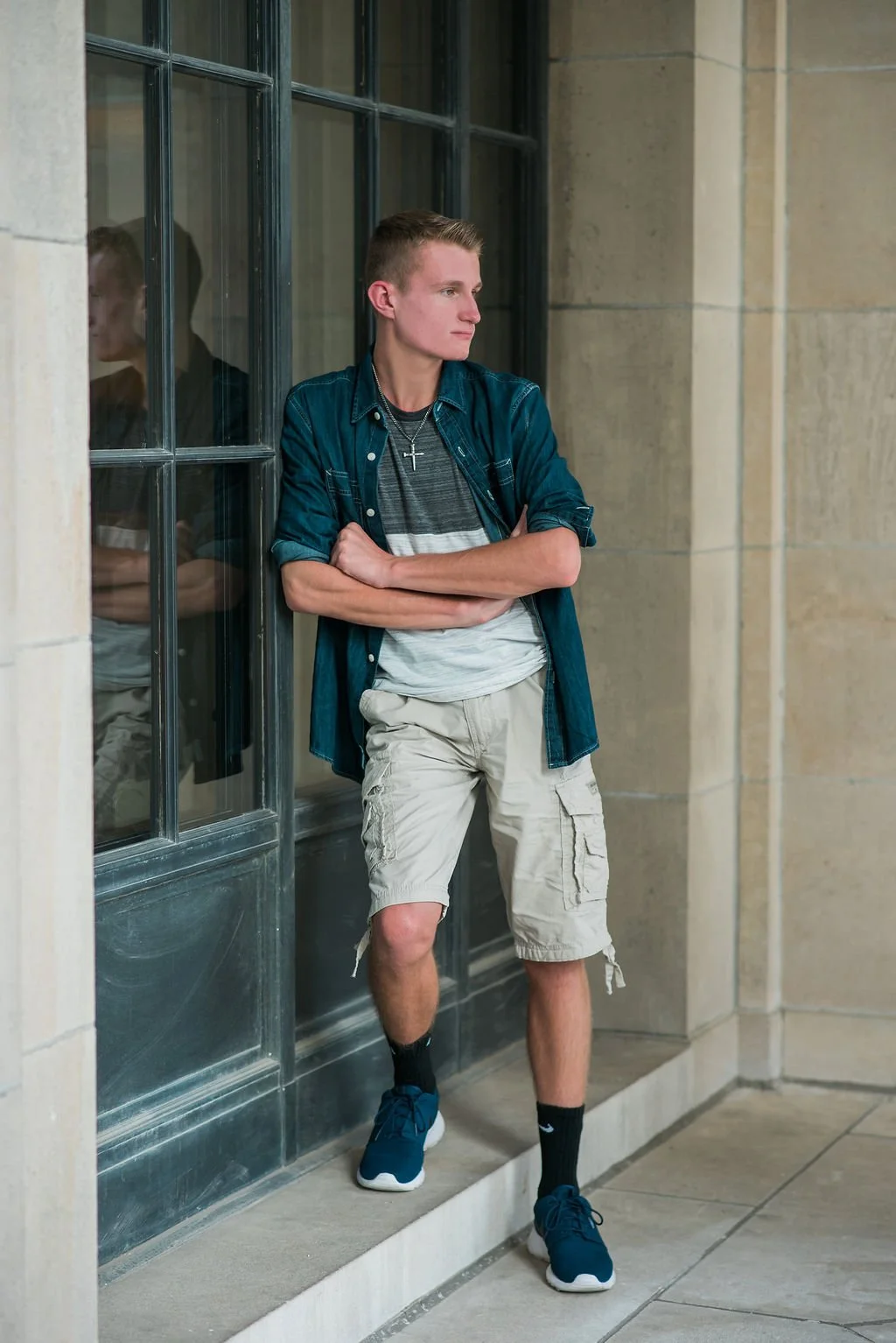 Young man with short hair and cross necklace leaning against a glass window with his arms crossed, wearing a blue denim shirt, striped t-shirt, beige cargo shorts, and blue sneakers, standing outside a building.