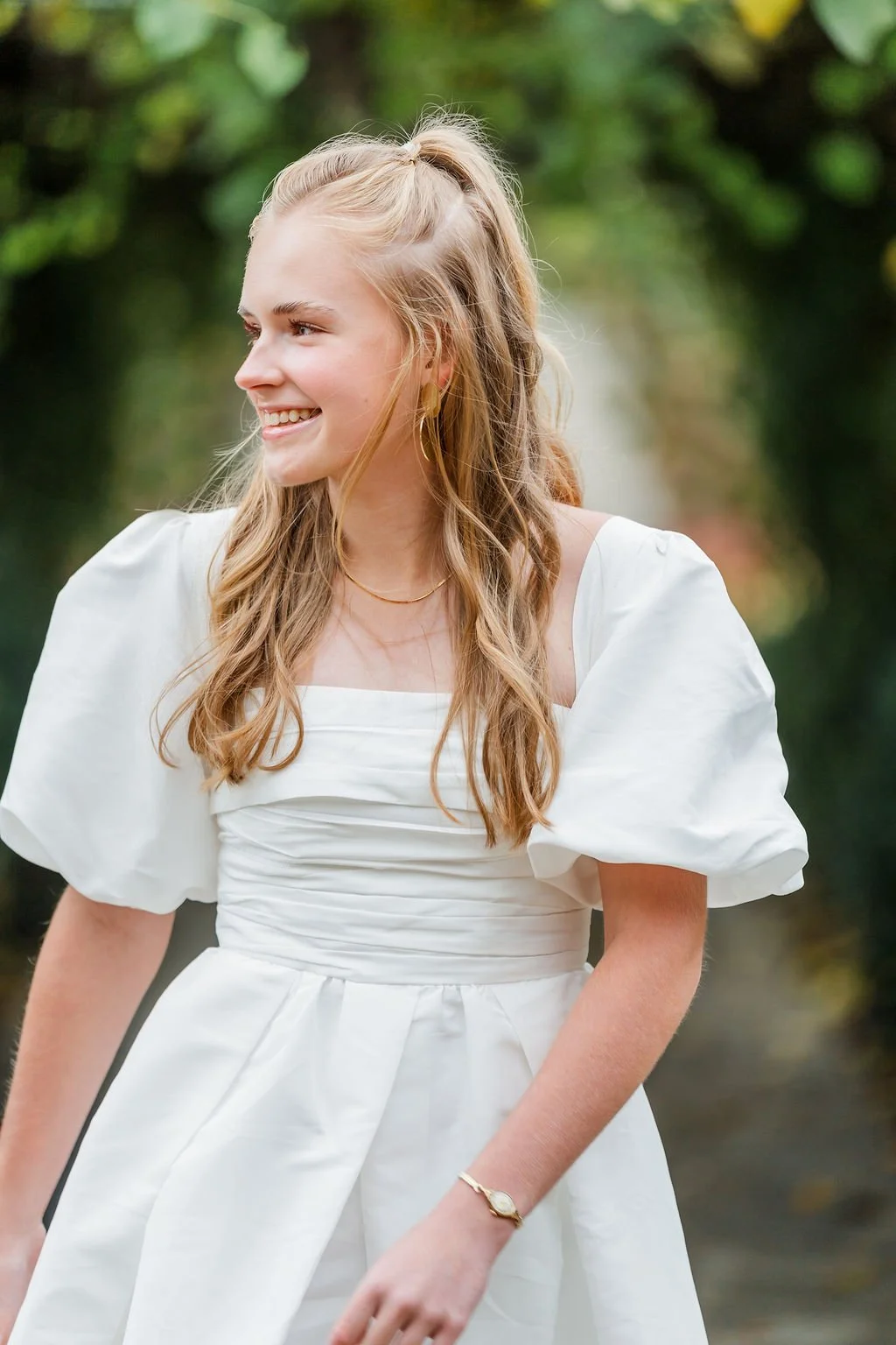 A young woman smiling outdoors, wearing a white dress with puff sleeves and gold jewelry, with greenery in the background.