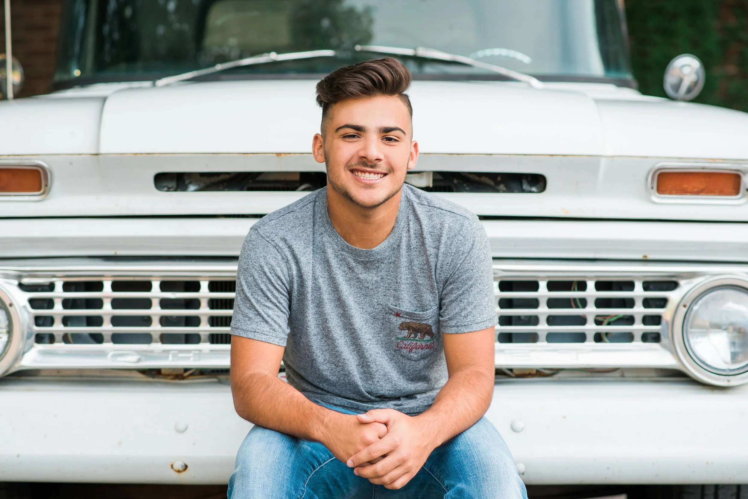 A young man with dark hair and a light beard smiling, sitting in front of a vintage white car, wearing a gray t-shirt and jeans.