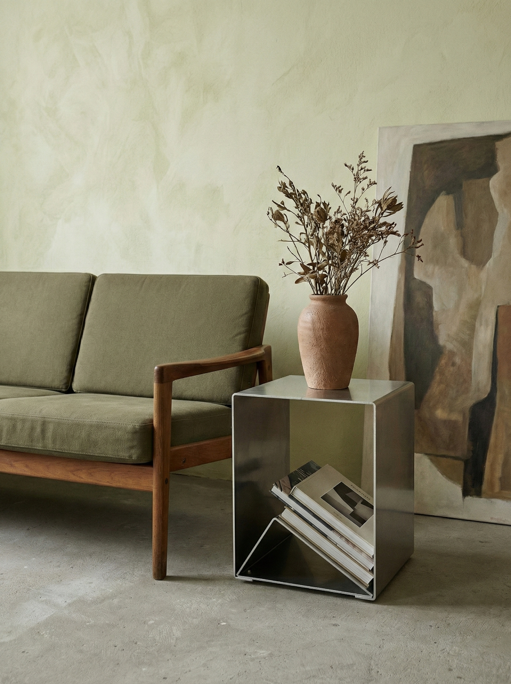 A green couch next to a aluminium side table holding a large clay vase with dried flowers. A framed abstract artwork leans against the wall behind the table. The floor is concrete, and the wall is painted a soft light green.