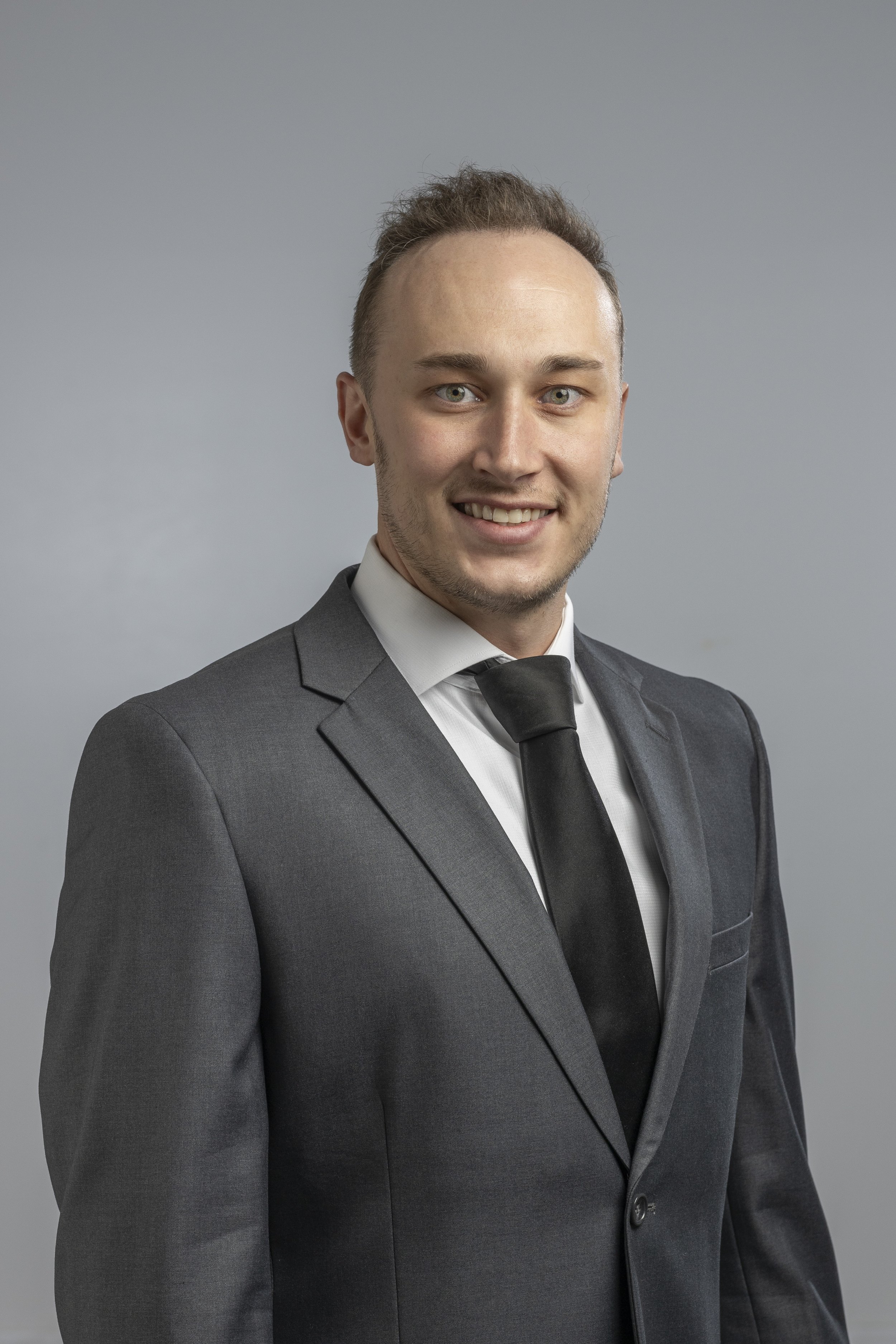 Professional portrait of a young man with light brown hair, blue eyes, and light facial hair, wearing a dark gray suit, white shirt, and black tie, against a plain gray background.