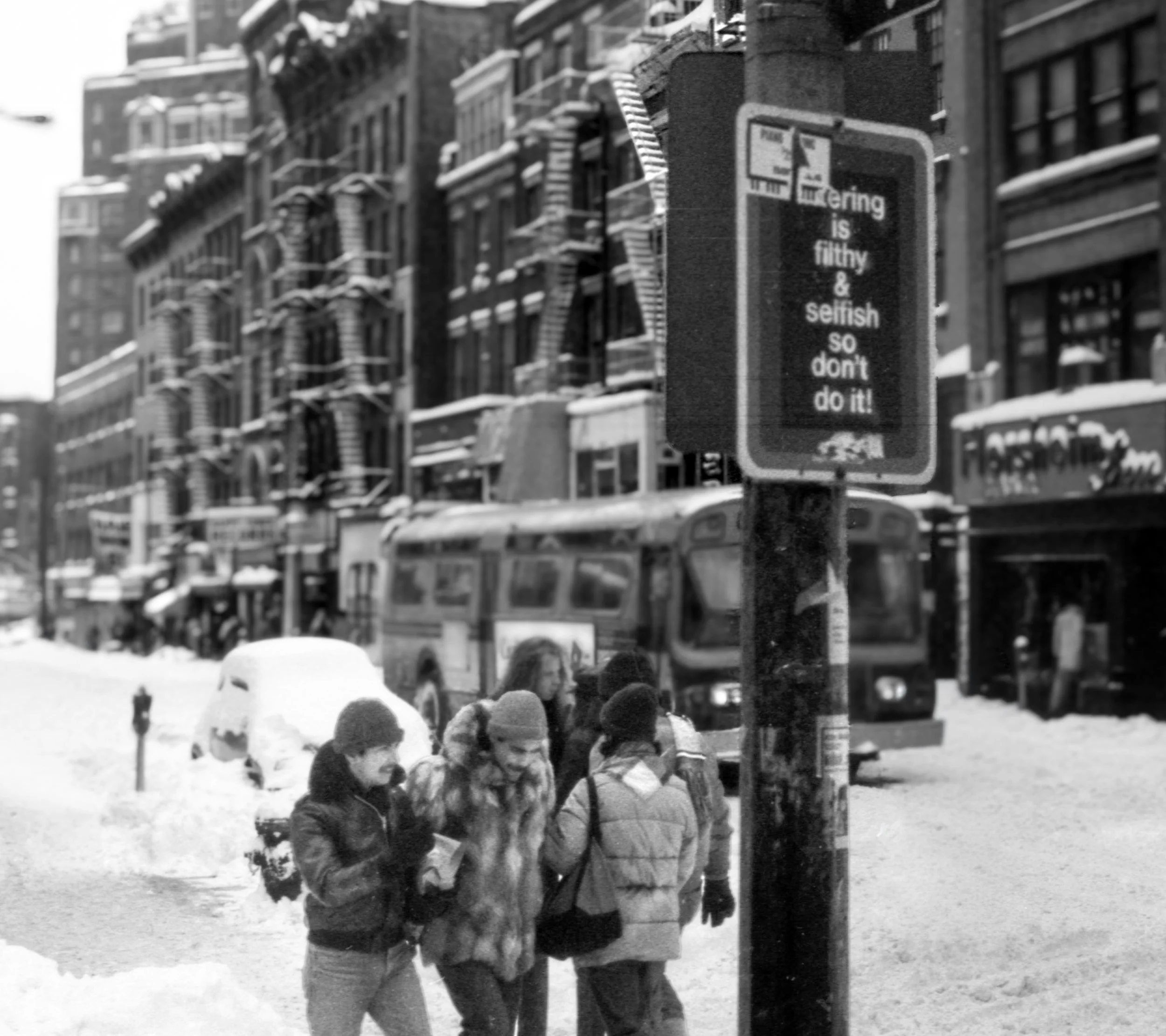 Black and white photo of a snowy city street with a group of people walking, a bus, and buildings in the background. A sign in the foreground reads, 'Littering is filthy & selfish so don’t do it!'.