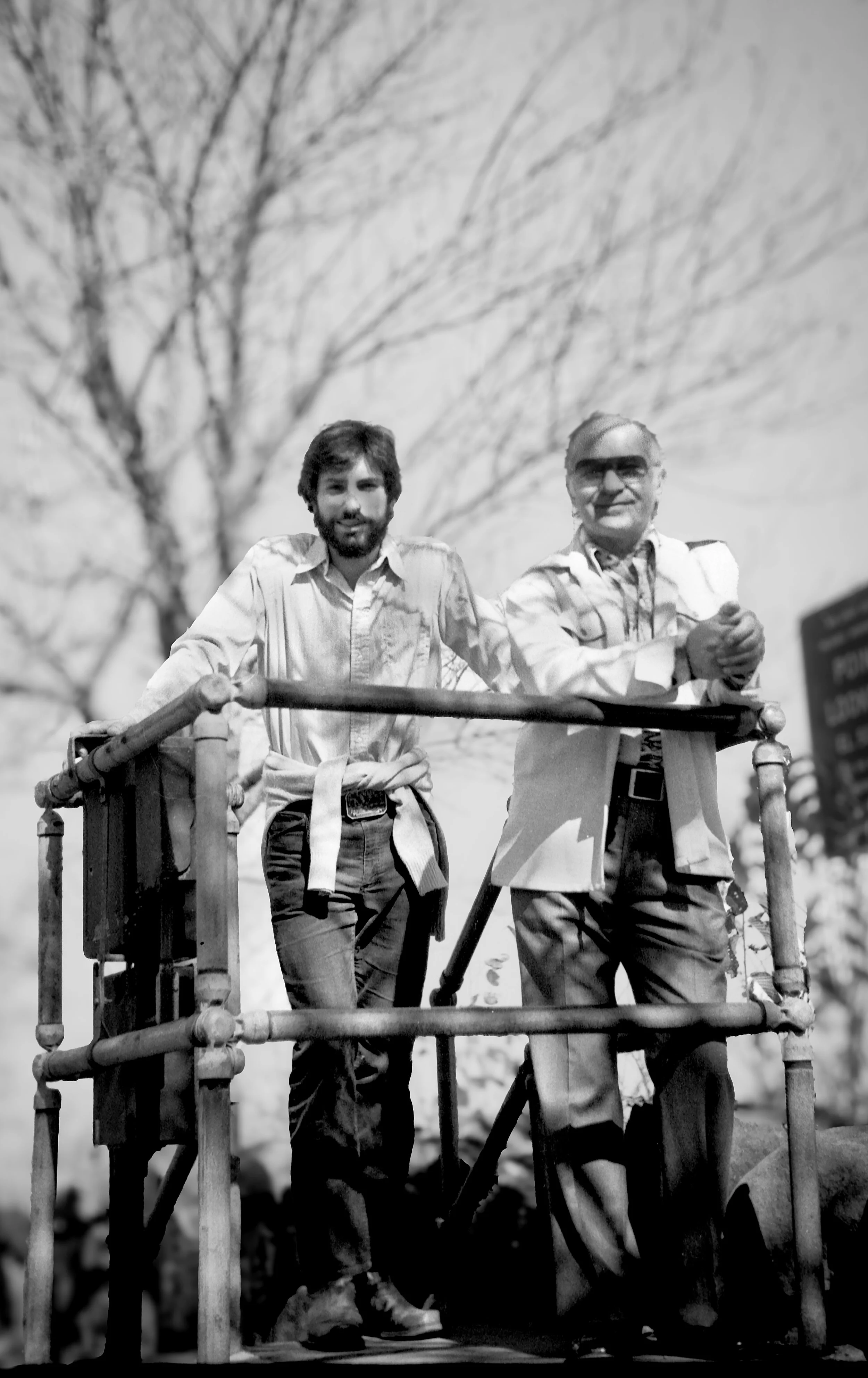 Black and white photo of two men standing on a platform outdoors, with a leafless tree in the background. They are leaning on the railing and looking at the camera.