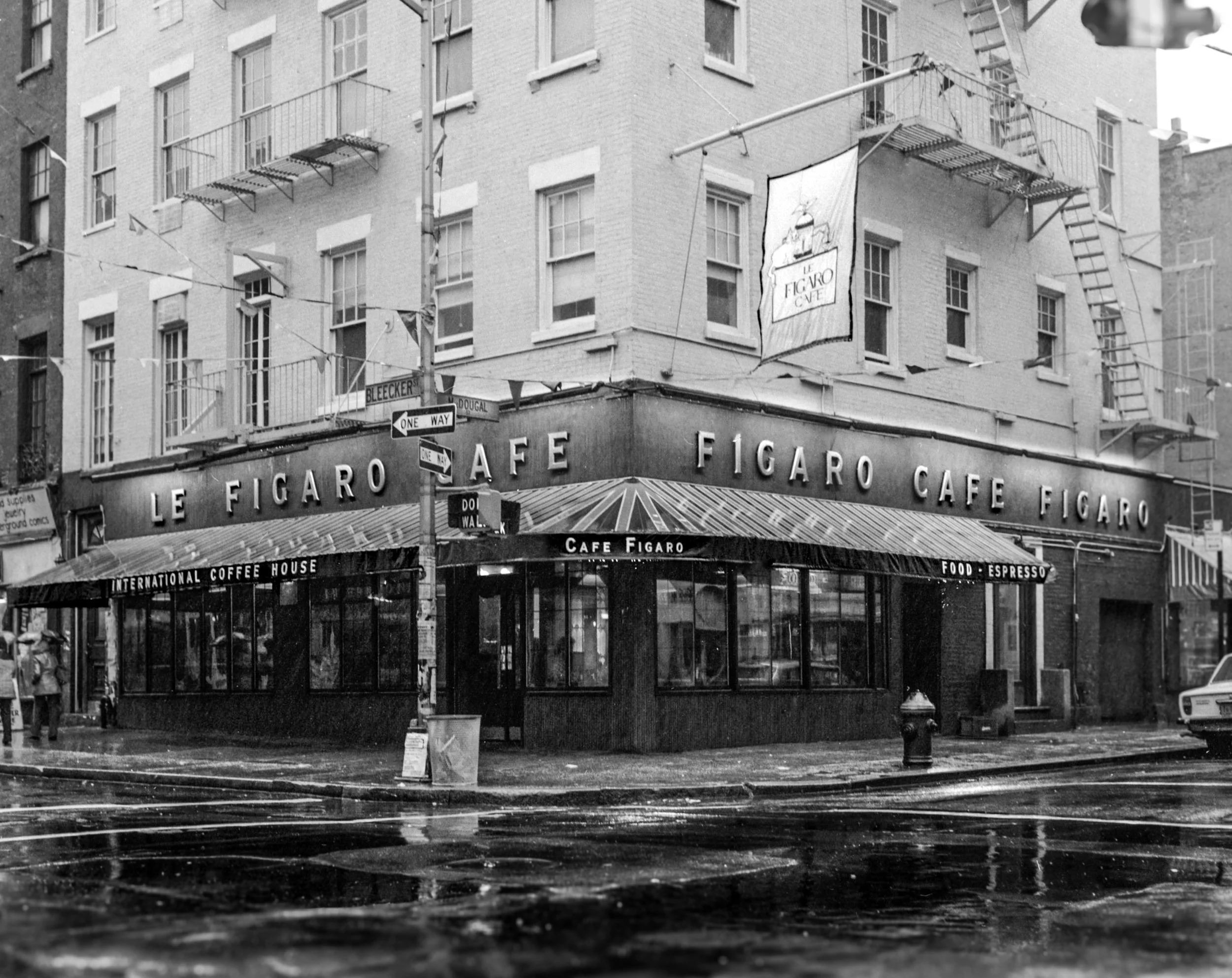 Black and white photo of Le Figaro Cafe on a street corner, with building signs, awnings, and a few pedestrians and cars in front.