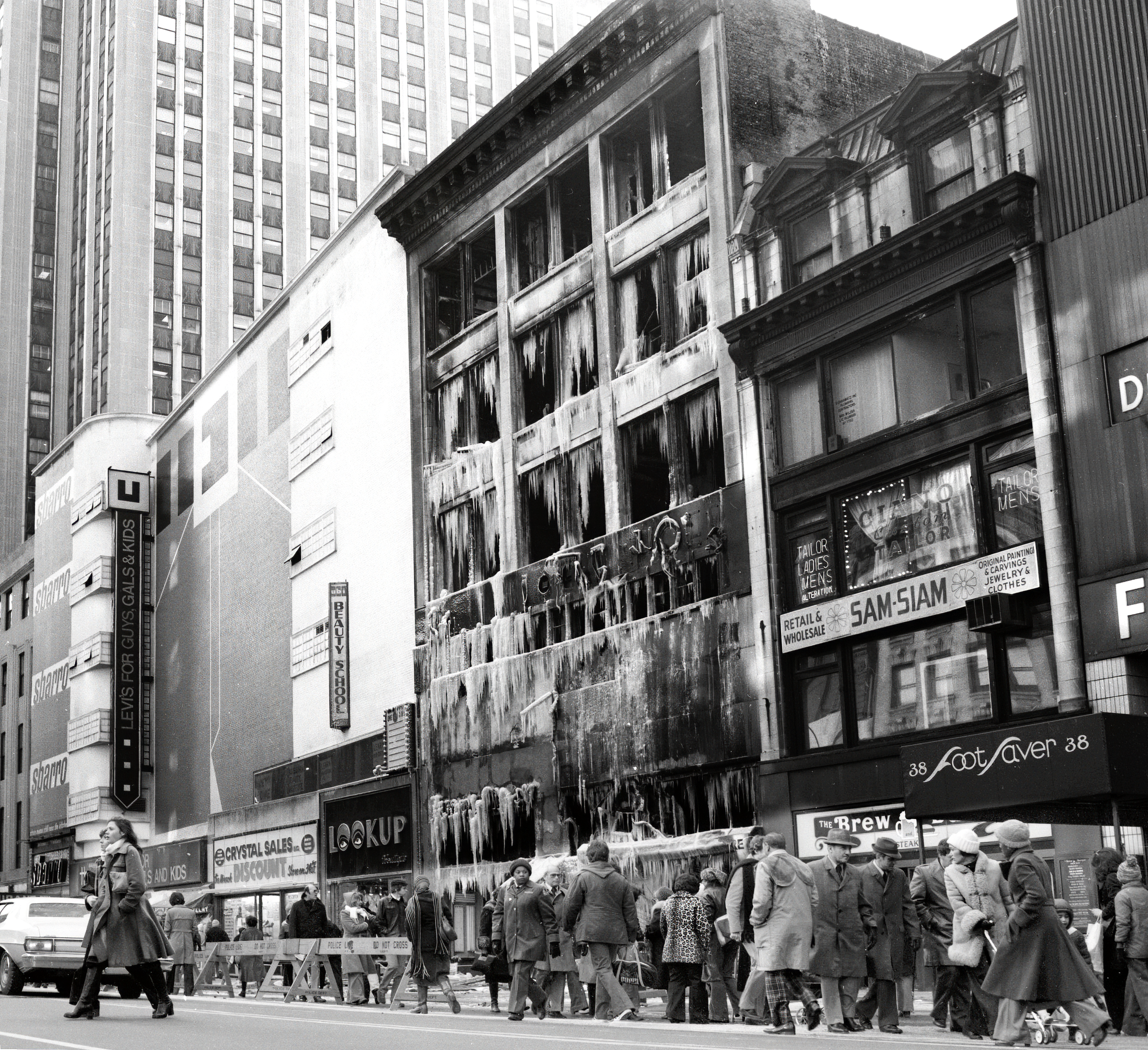 A city street scene showing a burned-out building with icicles hanging from it, surrounded by pedestrians and other buildings.