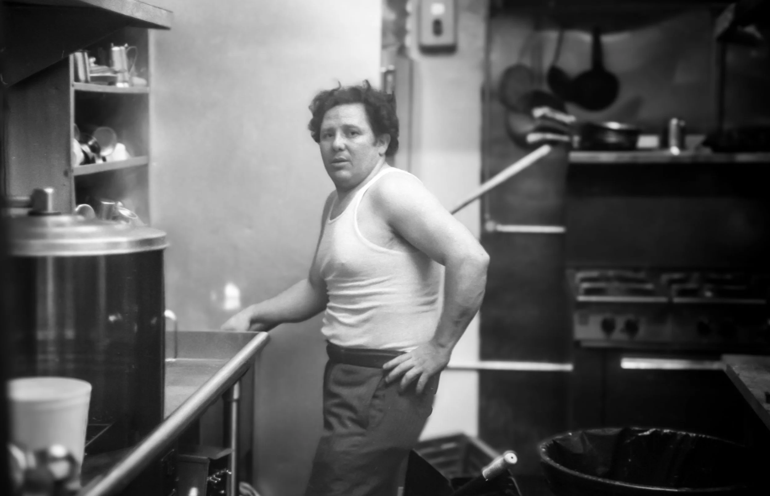 A man with curly hair wearing a white tank top and dark pants, standing in a kitchen, looking at the camera with a surprised or confused expression.