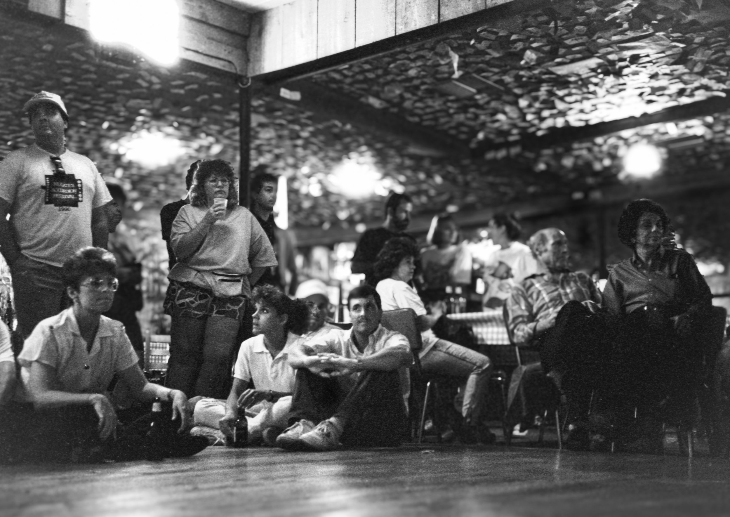 Black and white photo of a group of people sitting and standing in a dimly lit indoor setting, possibly a bar or restaurant, with a textured ceiling and some overhead lighting.
