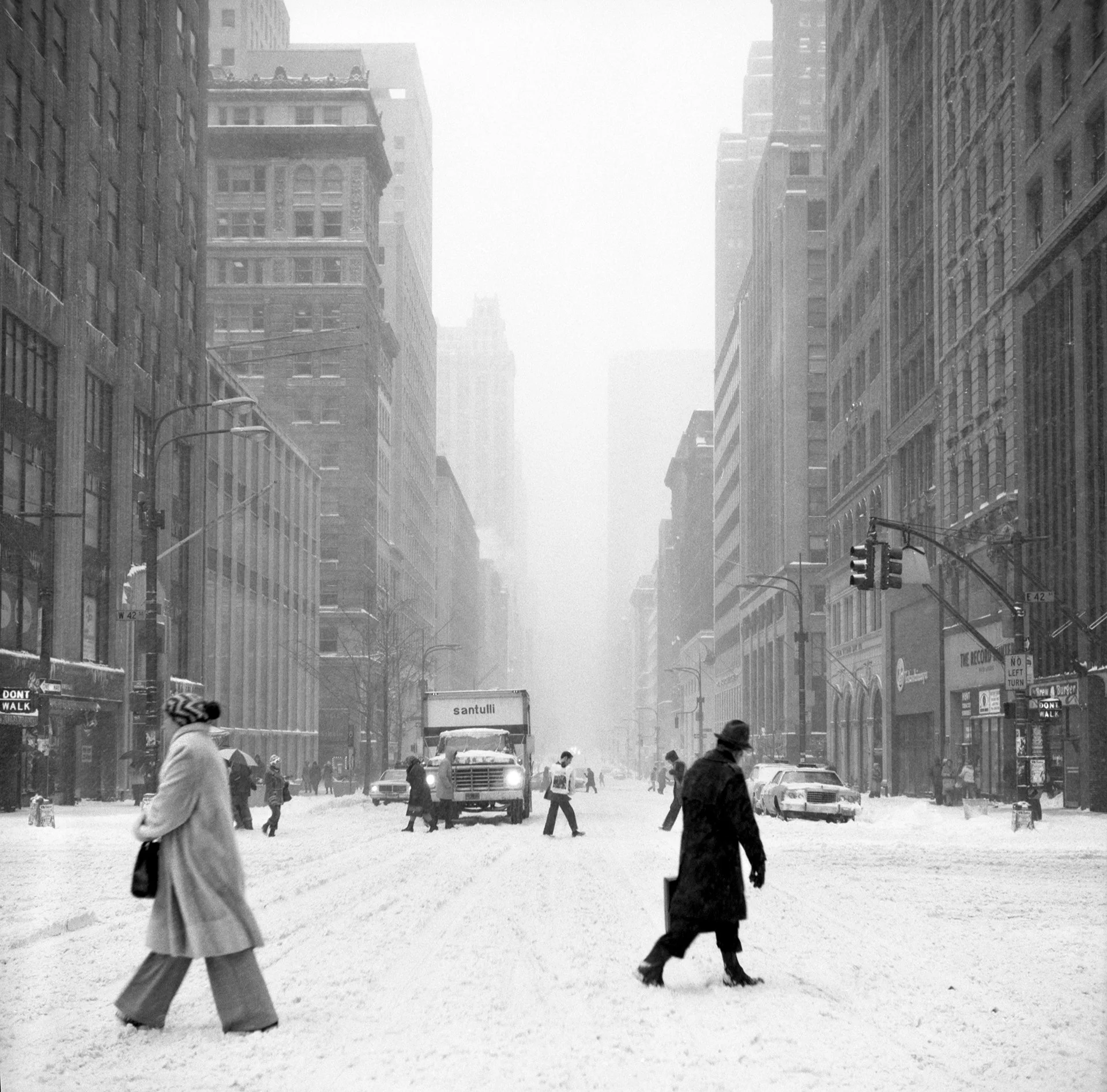 Black and white photo of a city street during a heavy snowstorm with pedestrians walking, some carrying umbrellas, and snow-covered cars and buildings lining the street.