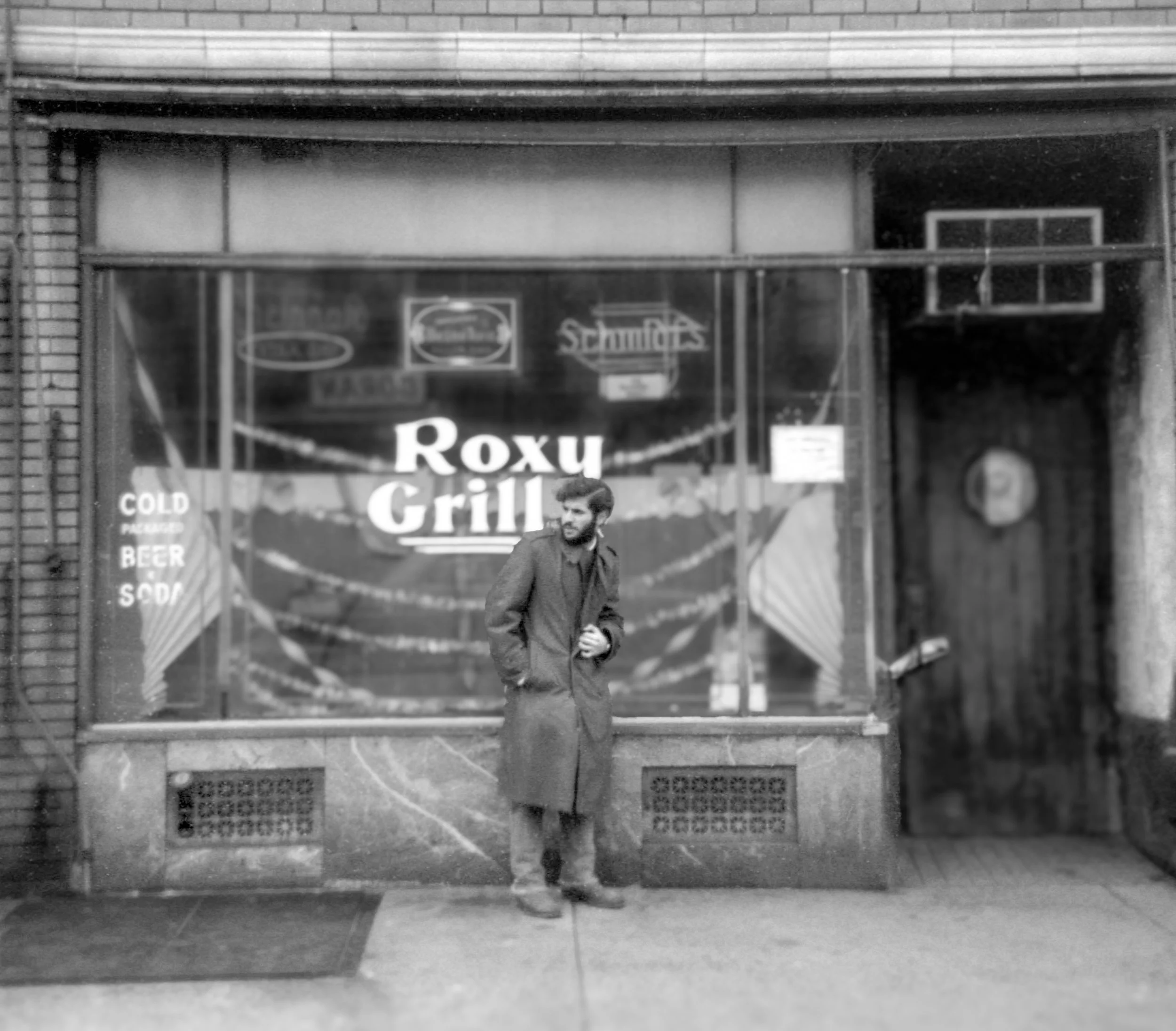 A man in a long coat standing outside a restaurant called Roxu Grill, with a large window displaying signs advertising cold packaged beer and soda.