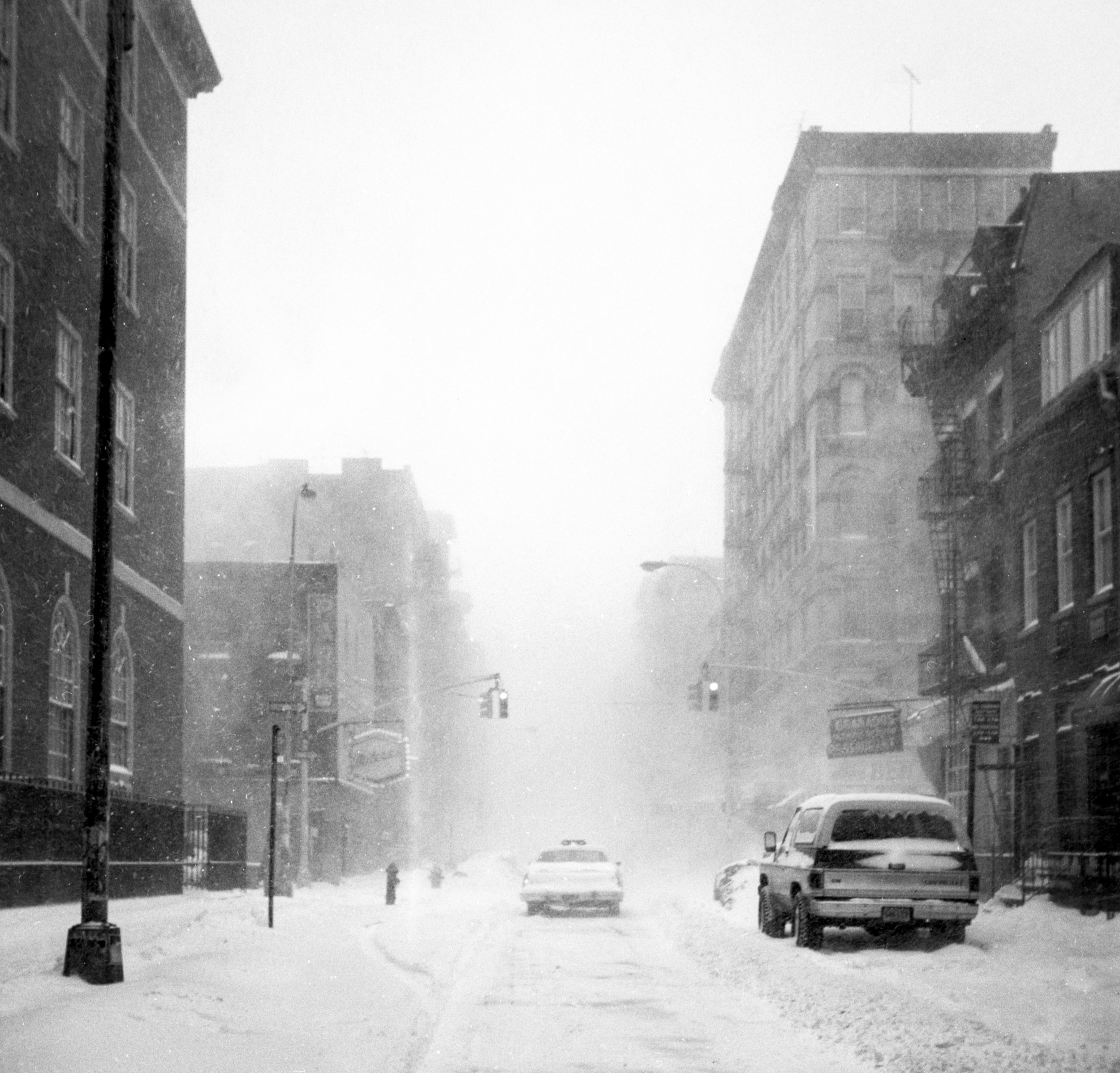 Snow-covered city street with parked cars, multi-story brick buildings, and streetlights, with snow falling and visibility reduced.