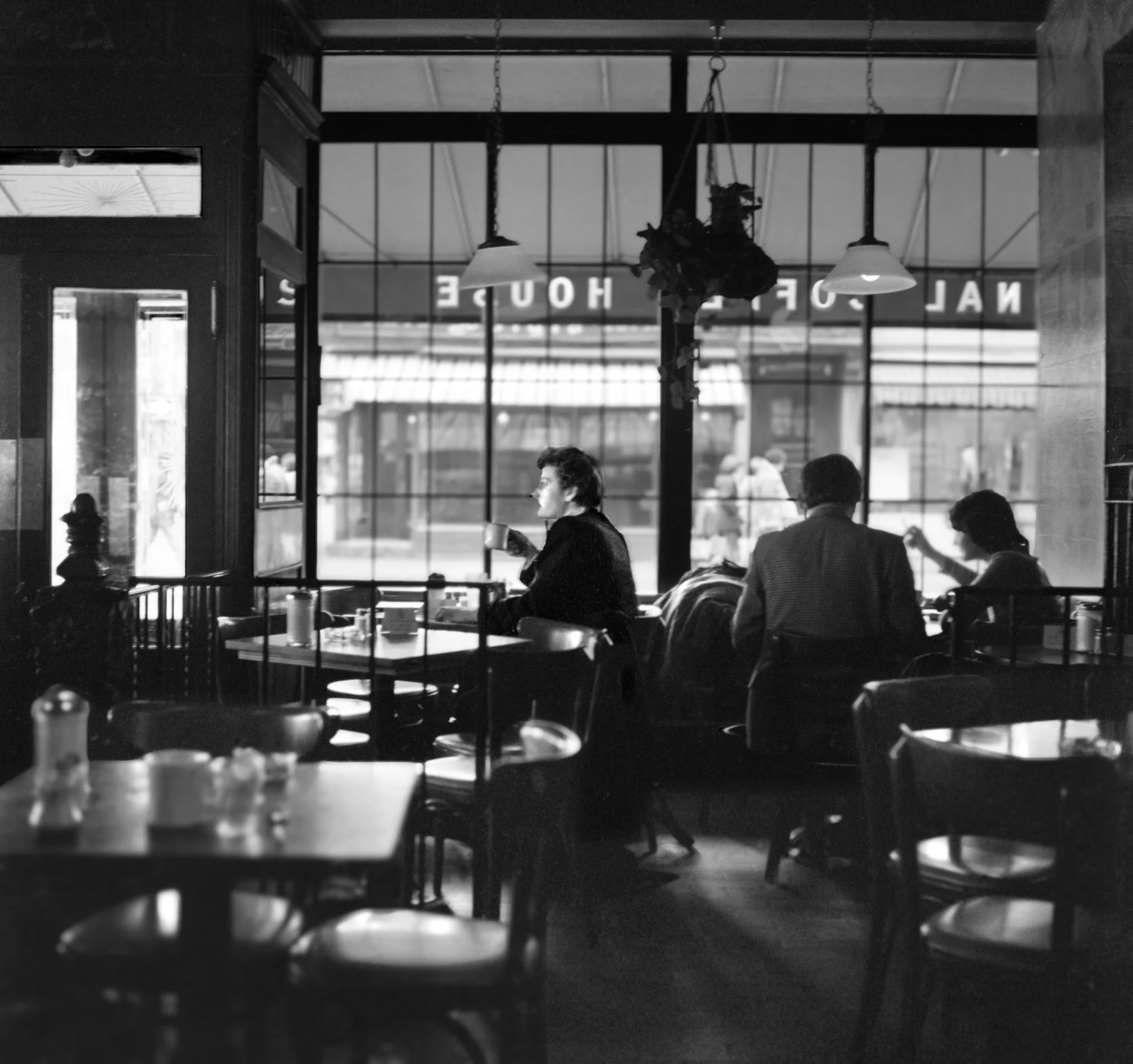 Black and white photo of a cozy cafe interior with three people sitting at tables near large windows. One person is drinking from a mug, another is working or reading, and the third person is seated, facing away from the camera. There are chairs and 