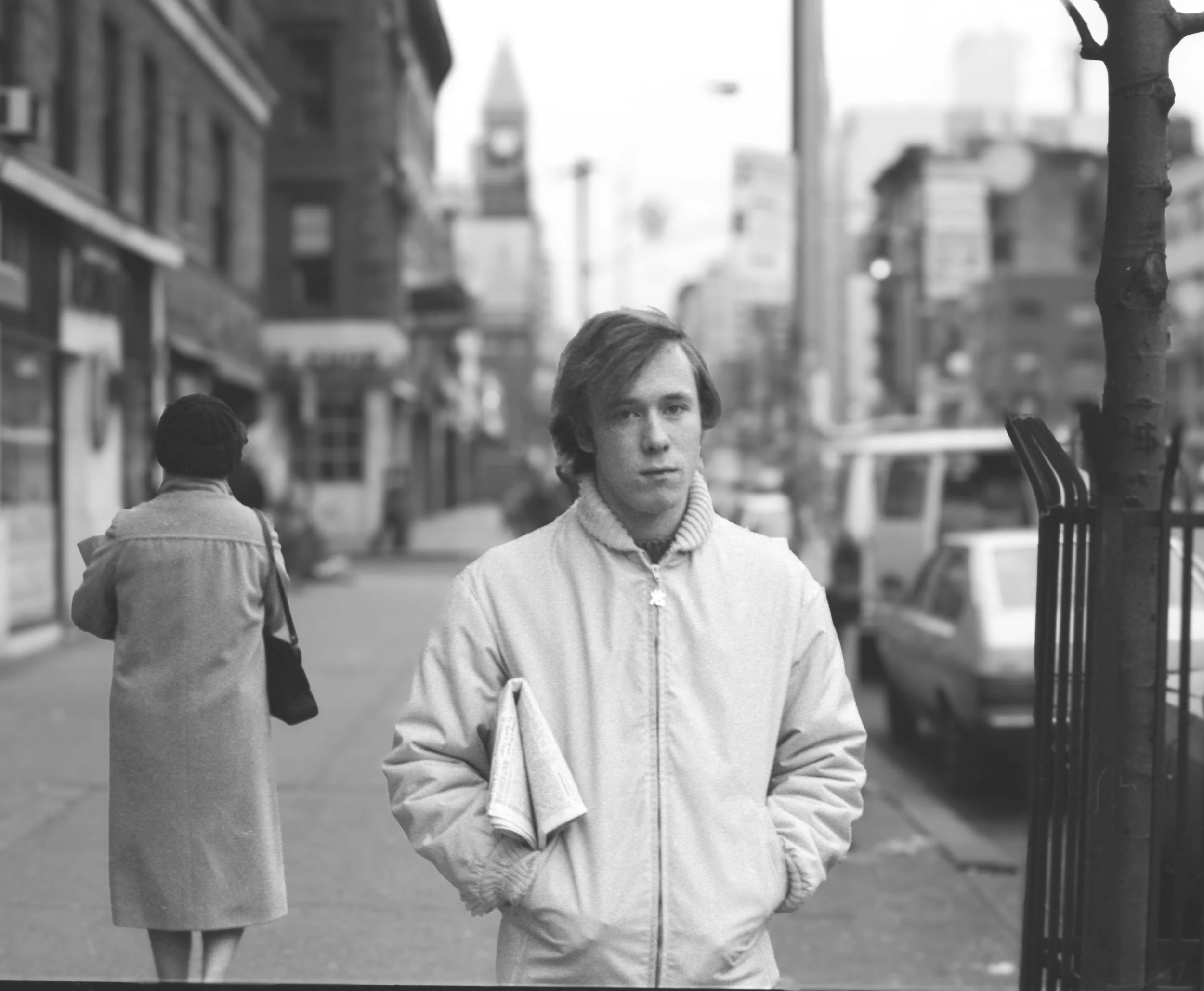 A black-and-white photo of a young man with medium-length hair standing on a busy city street, wearing a light-colored jacket, with a newspaper tucked into his pocket, looking into the camera. In the background, a woman with a coat and handbag is wal