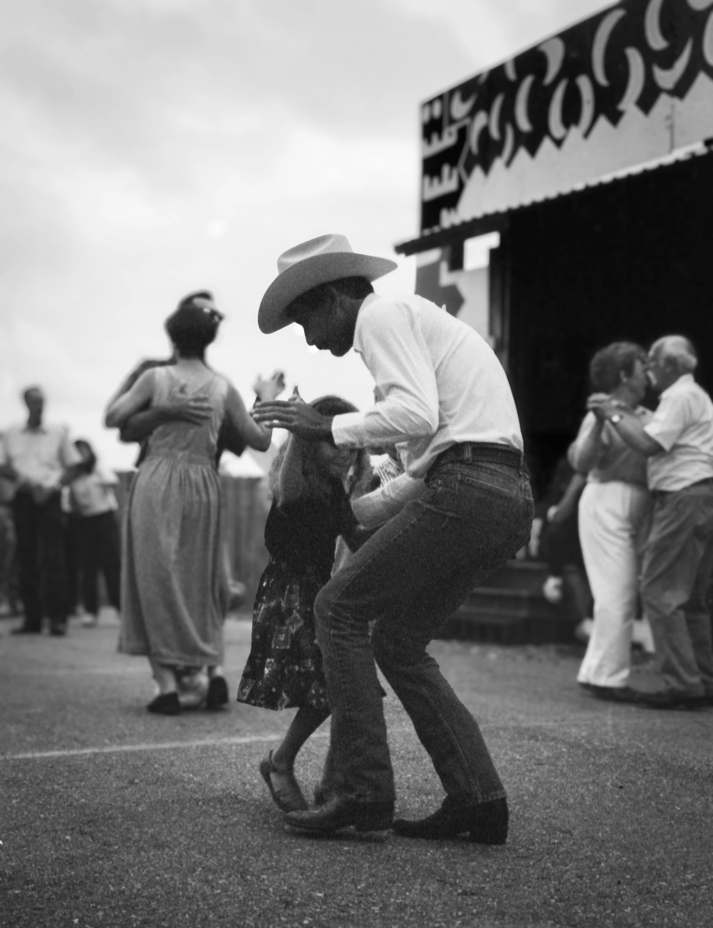 A black and white photo of people dancing outdoors, including a man wearing a cowboy hat and a woman in a floral dress embracing with others dancing nearby.
