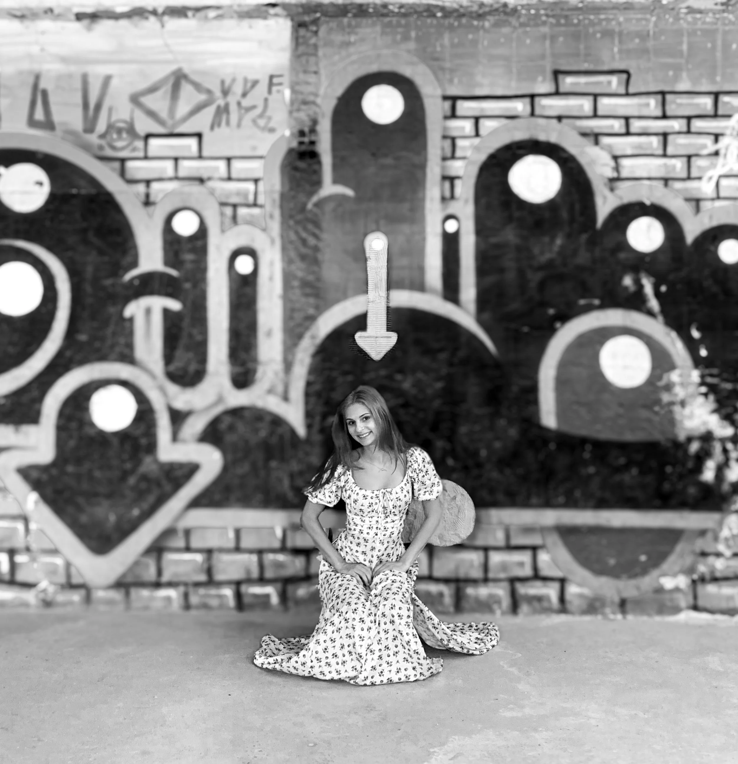 A young woman in a floral dress kneeling in front of a graffiti mural with arrow and circle designs on a brick wall.