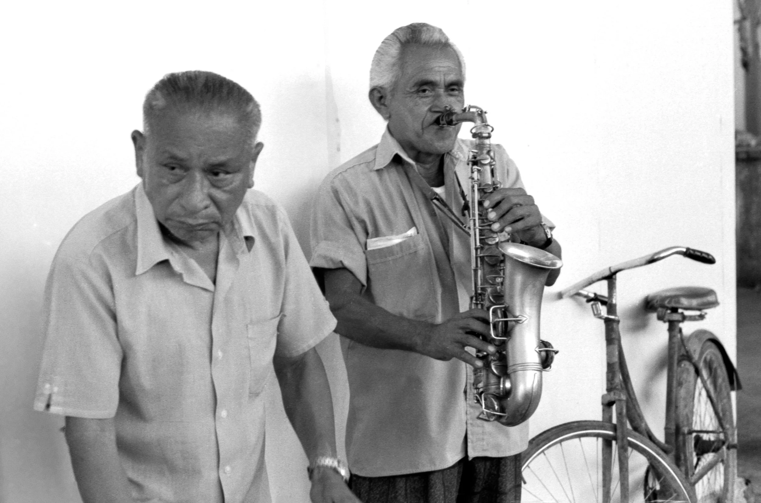 Two elderly men, one of whom is playing a saxophone, standing next to a vintage bicycle against a plain wall.