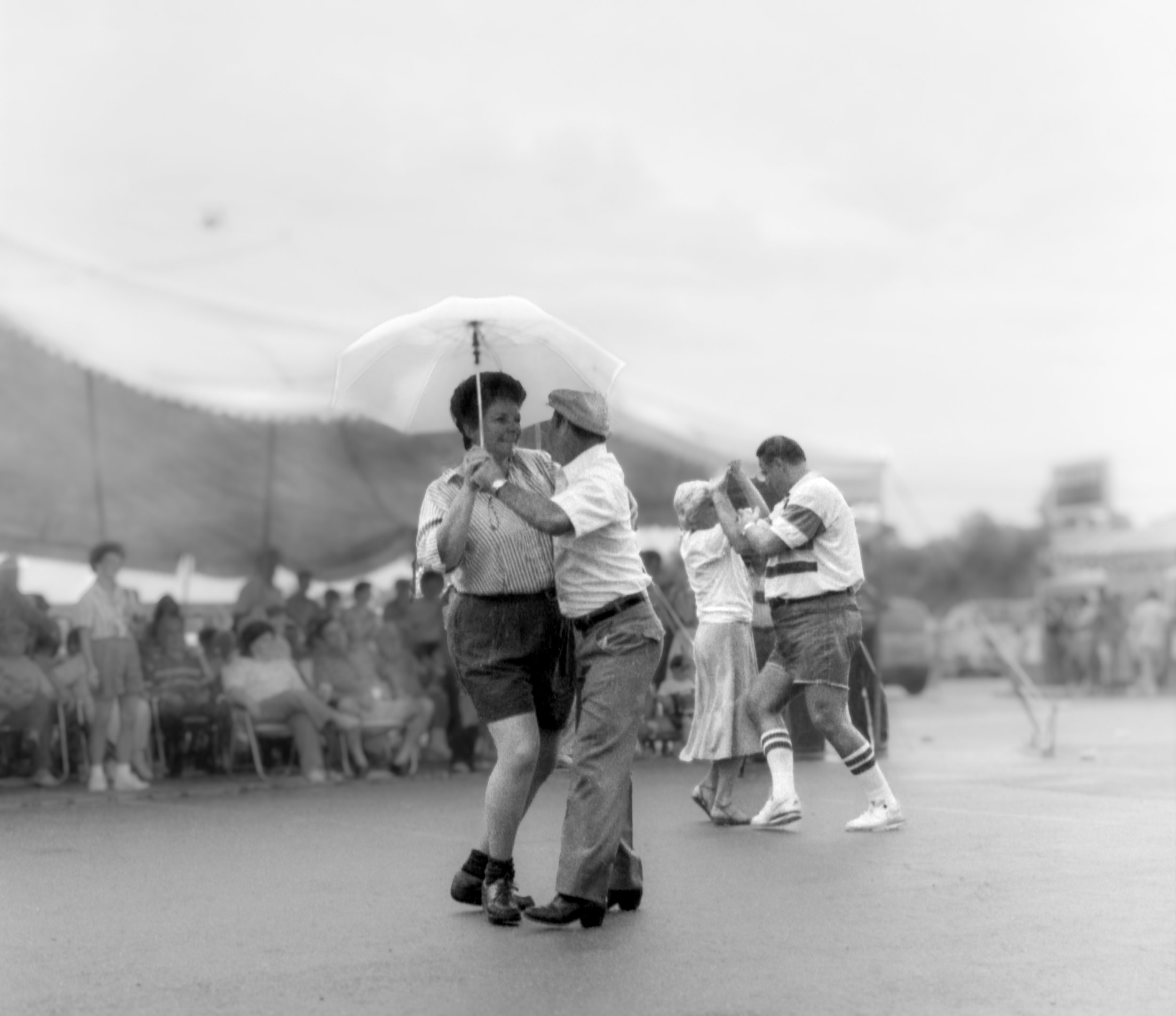 Black and white photo of a dance couple under a large umbrella, with other couples dancing and a crowd sitting on chairs in the background.