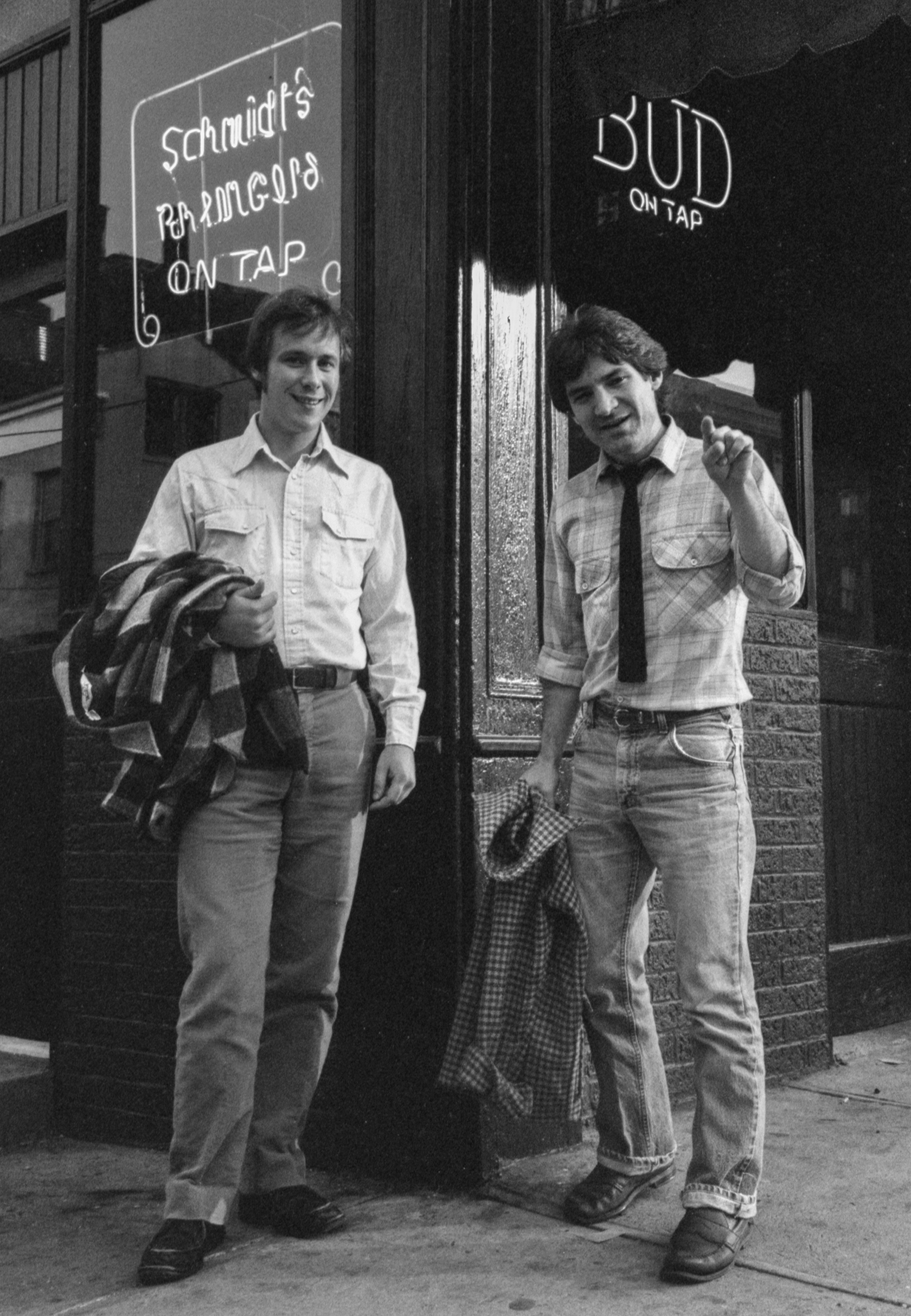 Two young men standing outside a restaurant, one holding a plaid shirt and the other holding a coat, smiling at the camera. The restaurant has neon signs advertising 'Schmaltz Ramen' and 'Bud on Tap'.