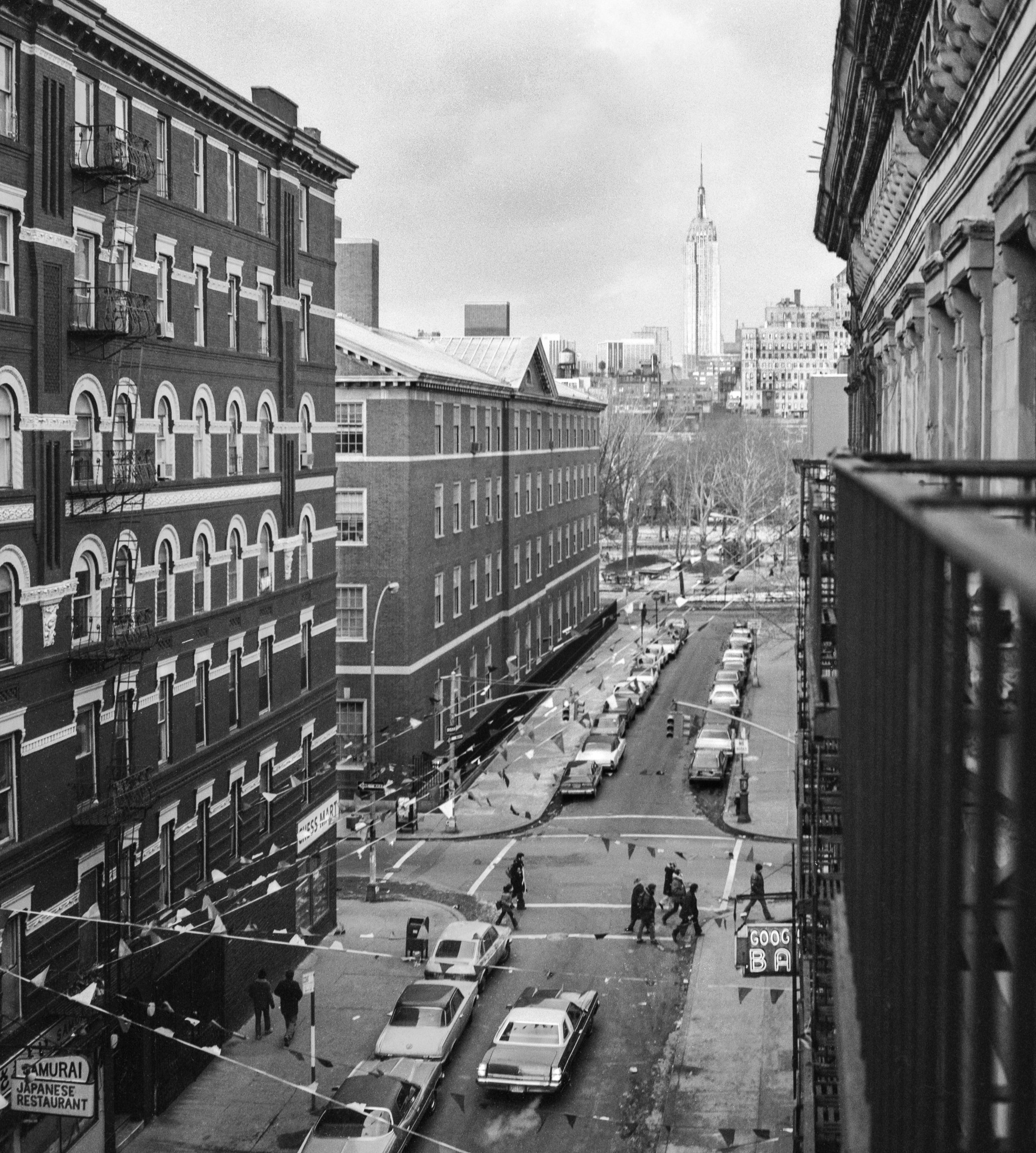 Black and white photograph of a city street viewed from a balcony, with parked cars, pedestrians crossing, multi-story buildings, and the Empire State Building in the background.