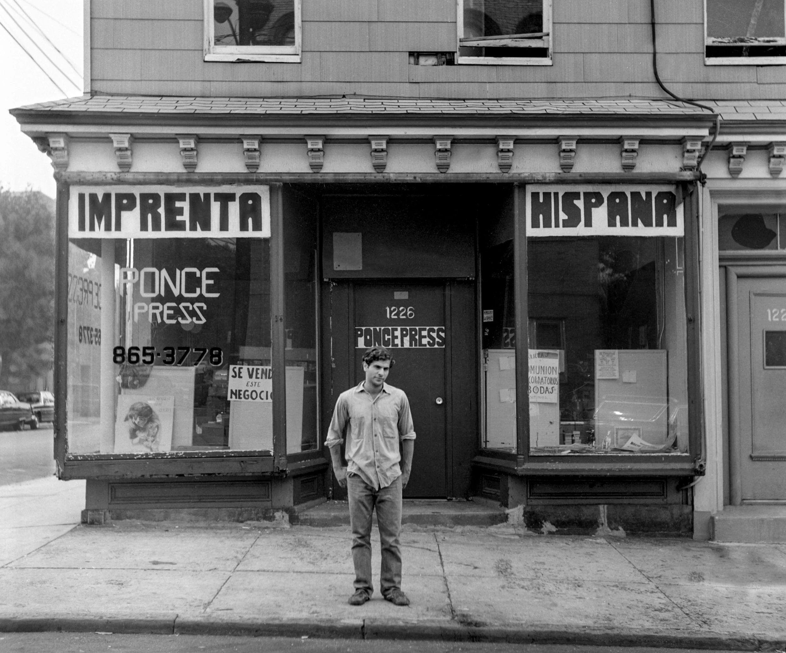 A man standing on a sidewalk in front of a storefront with large windows and signs that read 'IMPRENTA HISPANA' and 'PONCE PRESS.' The storefront appears old, with visible signs of wear and tear.