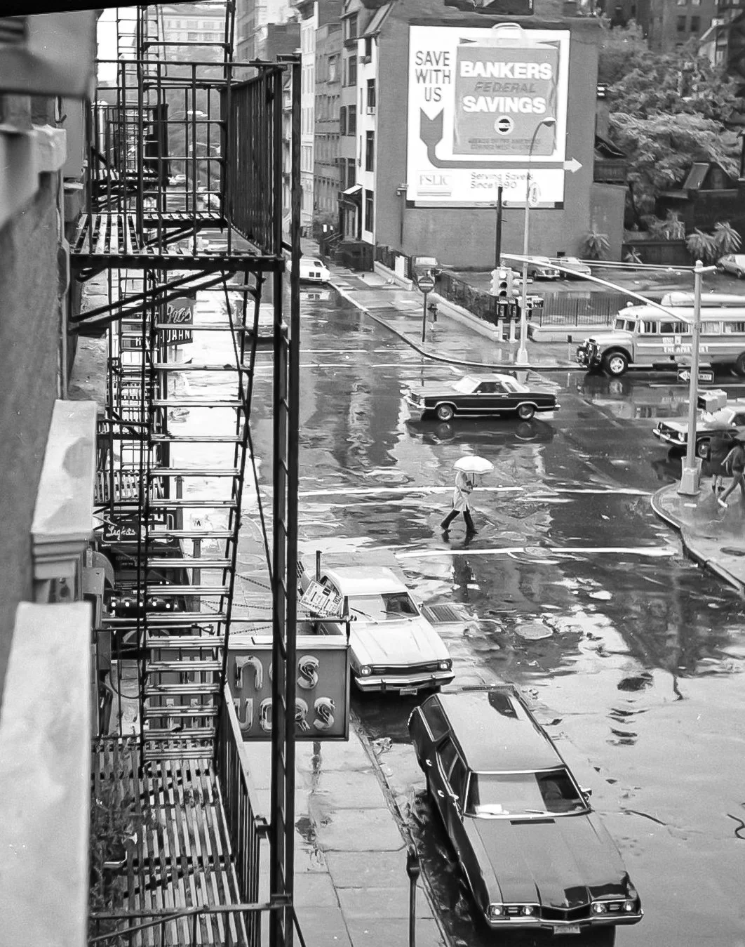 A rainy city street scene in black and white. There is a person walking with an umbrella across a wet crosswalk, several parked cars, and a bus. Tall buildings and a billboard advertising bank savings are visible in the background.