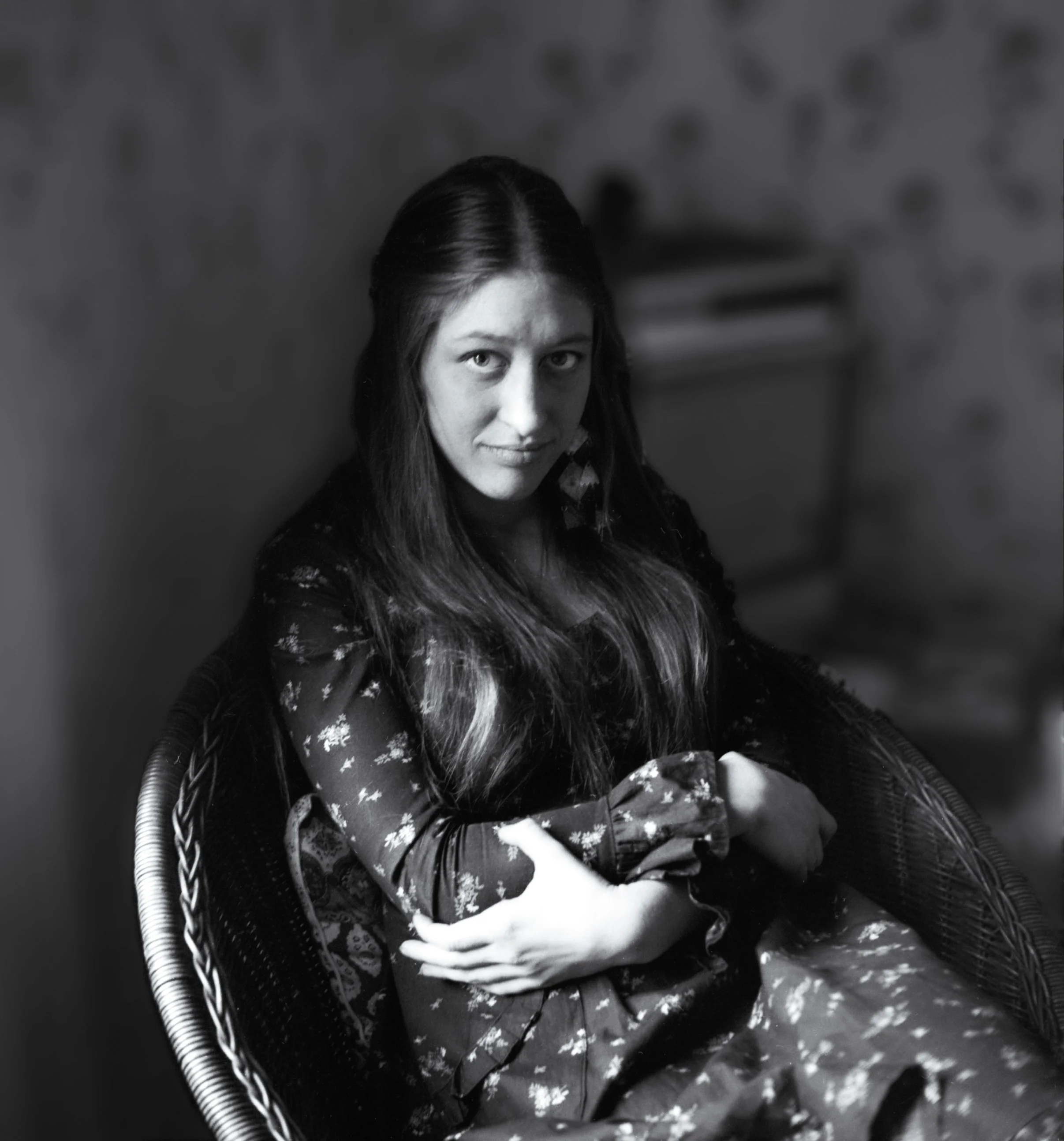 A woman with long dark hair sitting in a wicker chair, looking at the camera with a slight smile. The background is blurred.