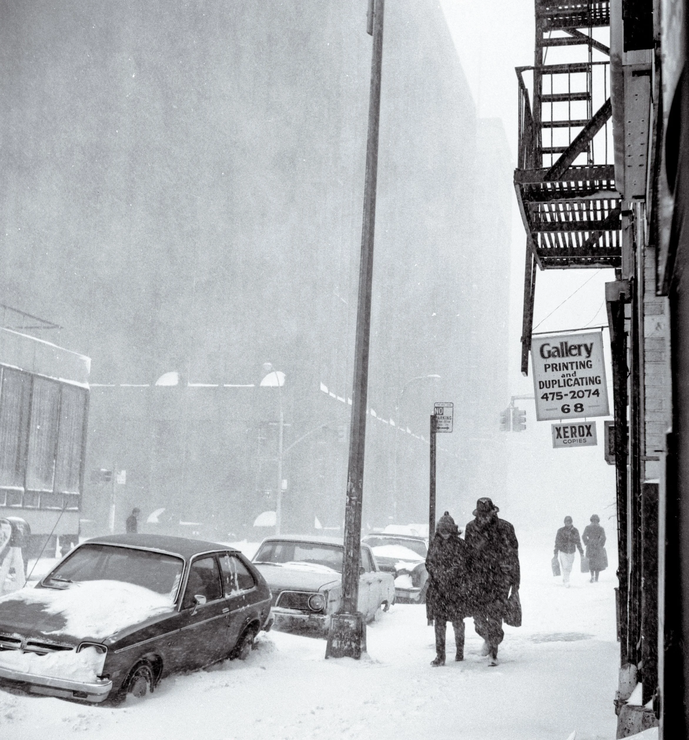 A black and white photo of a snowy street scene with parked cars, a storefront sign for a printing and duplicating business, and four people walking in the snow.