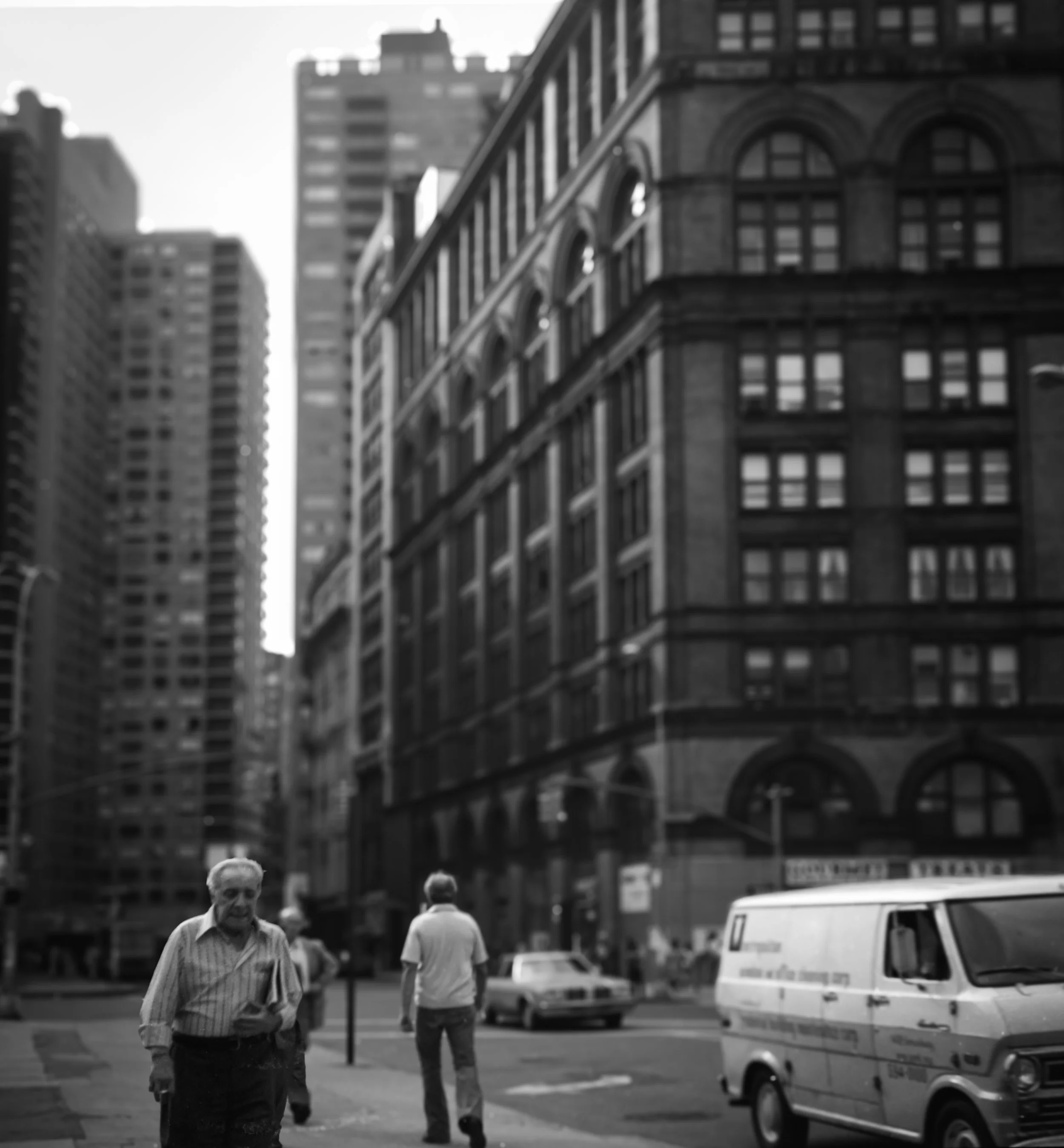 Black and white photo of a city street with tall buildings in the background. Pedestrians walk on the sidewalk, including an older man in the foreground, and a van is parked on the street.