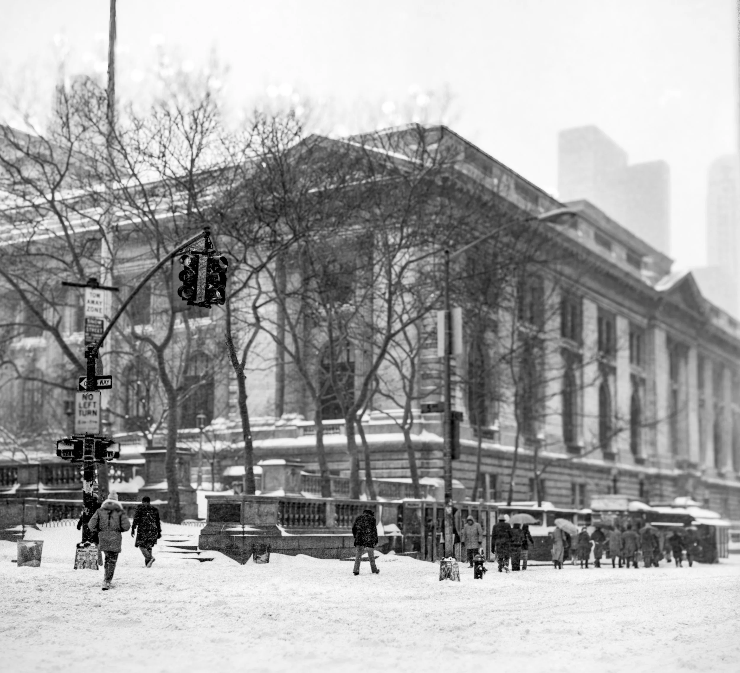 Black and white photo of people walking on a snowy city street, with a large historical building with columns and trees in the background, and traffic lights hanging overhead.