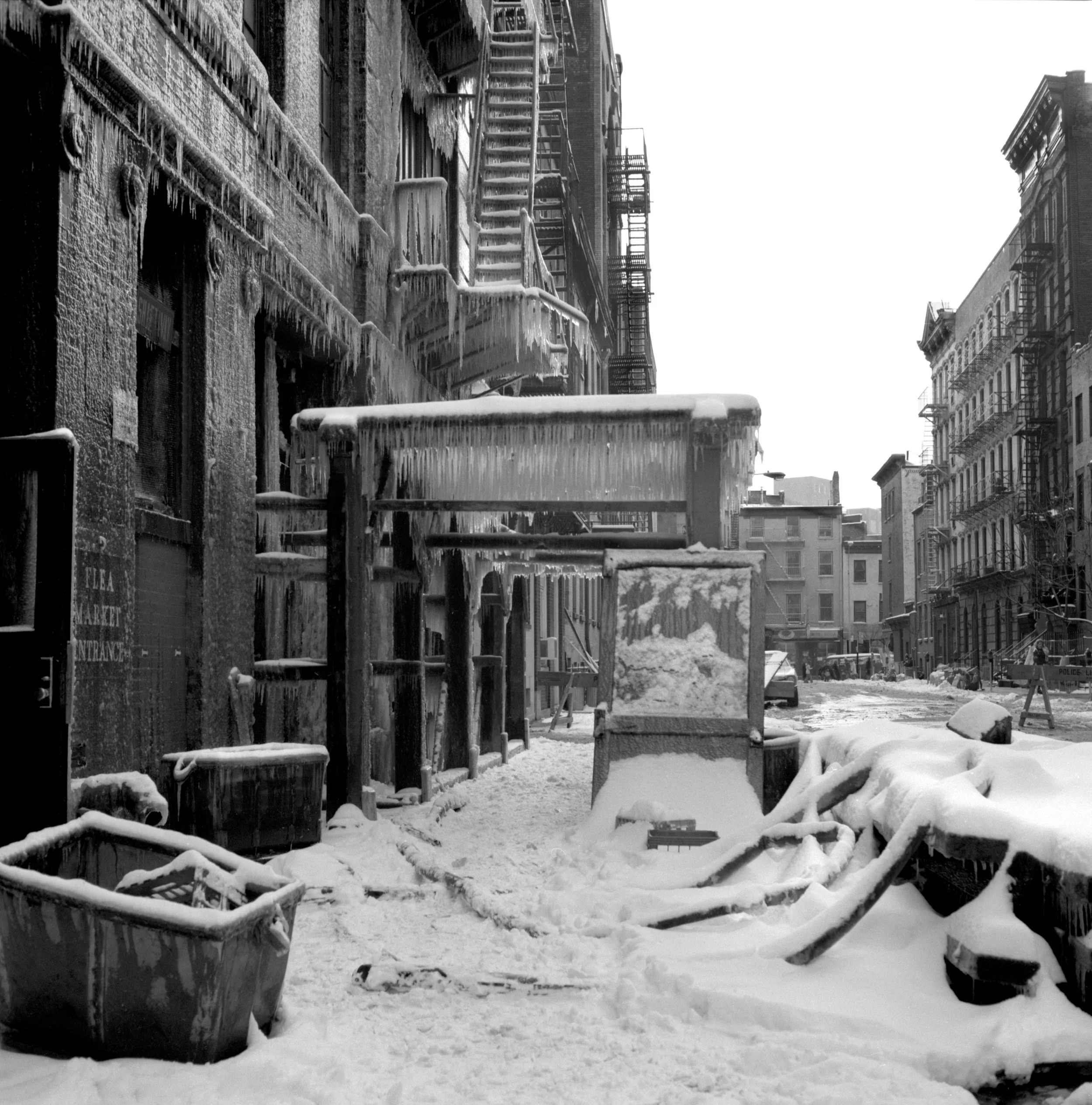 Snow-covered city street with frozen fire escapes, parked cars, and scattered debris.