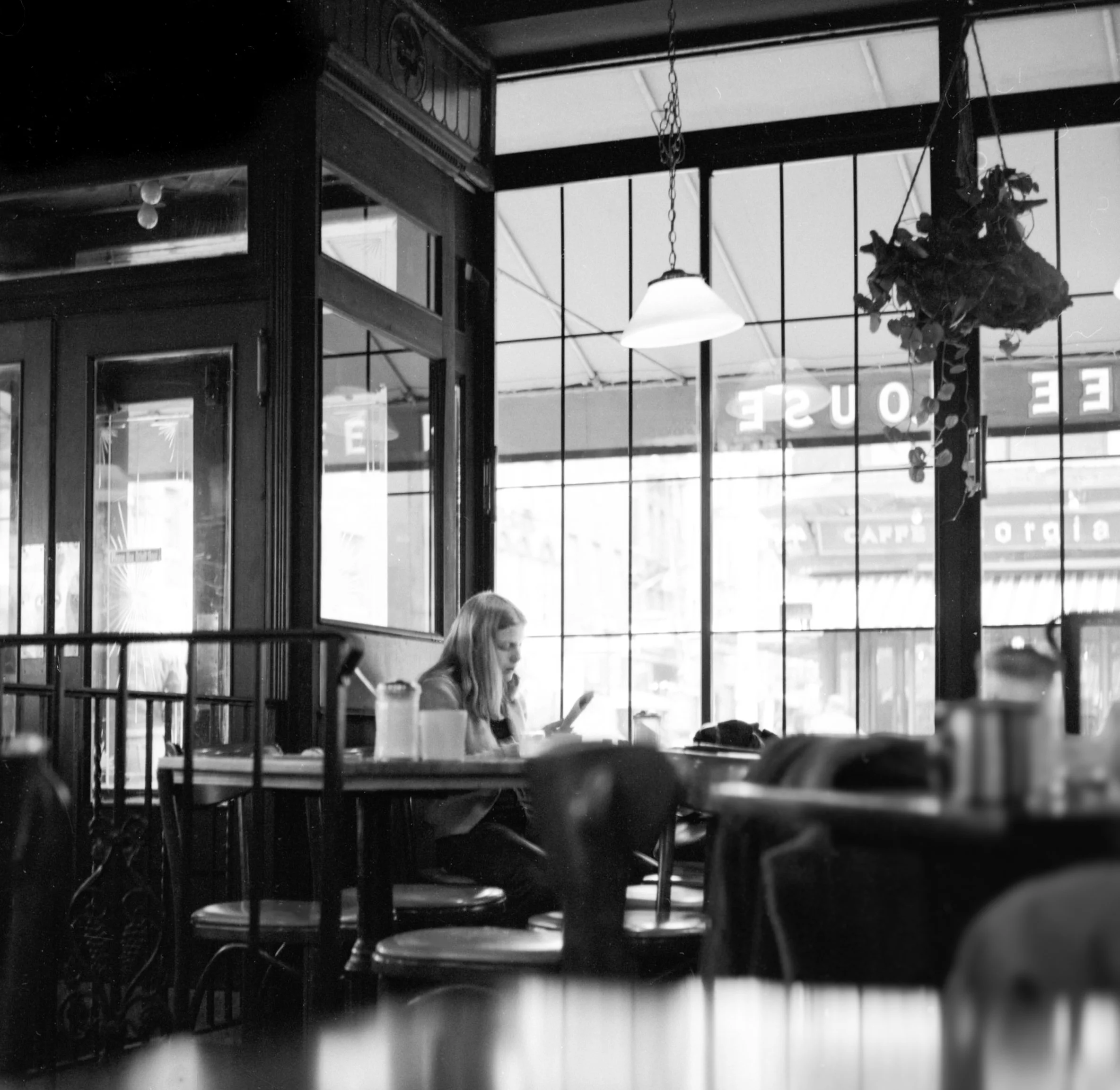 A woman sitting alone at a table in a cafe, looking at her phone, inside a space with large windows and hanging lights.
