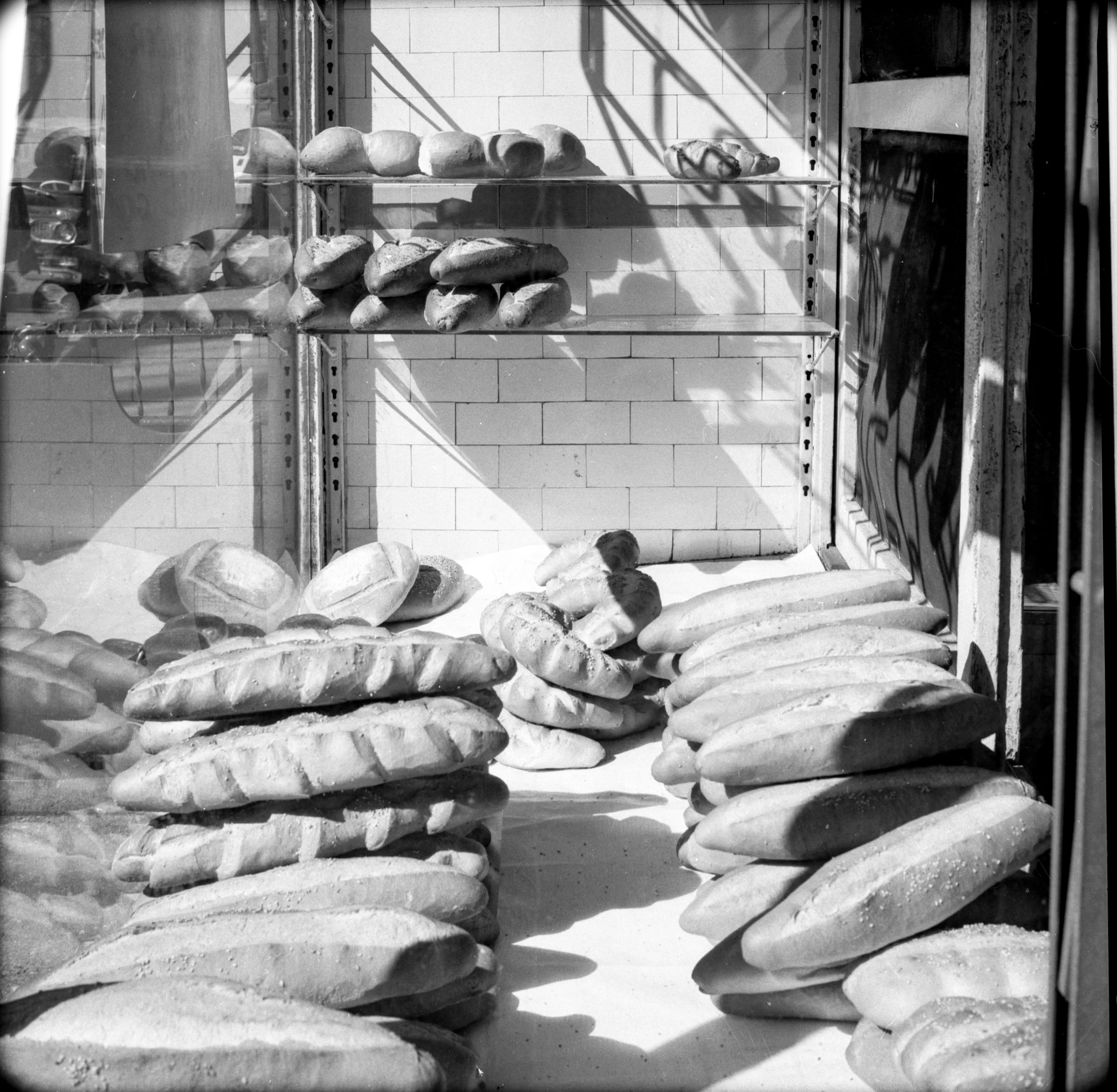 Display window with loaves of bread and baguettes stacked on shelves and on the floor, with shadows and reflections visible.