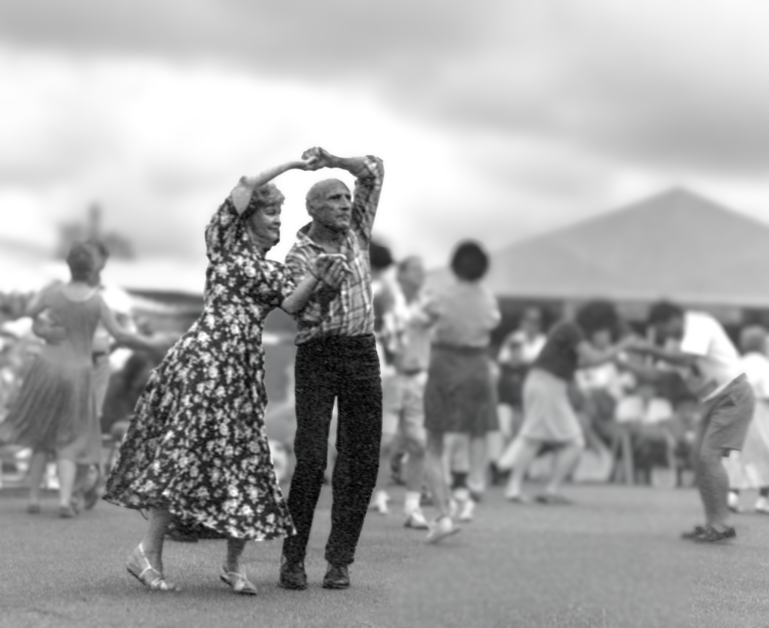 An elderly couple dancing hand-in-hand at a crowded outdoor event with tents and people in the background.