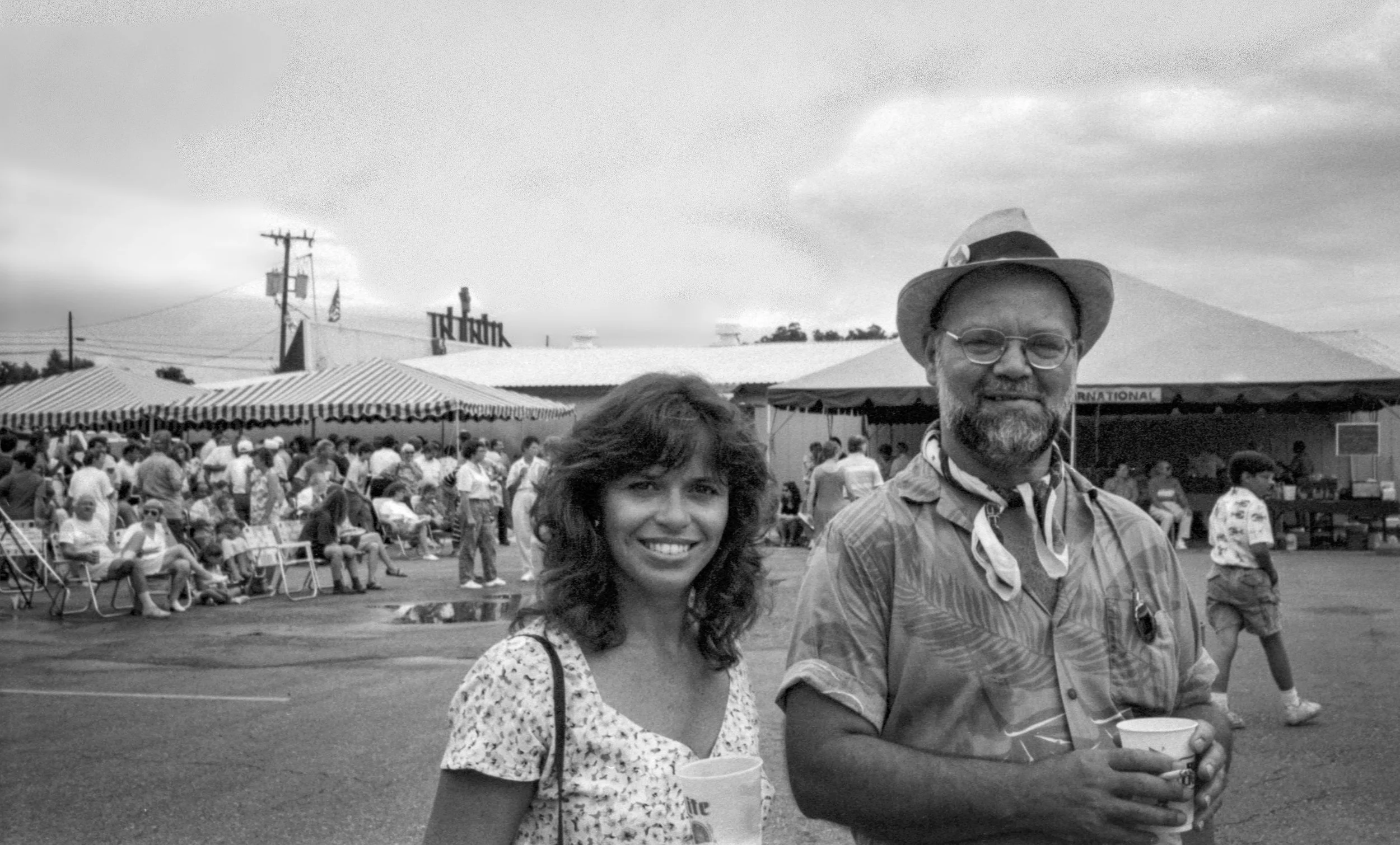 A black and white photo of a woman and a man smiling and holding cups at an outdoor event, with a large crowd and tents in the background.
