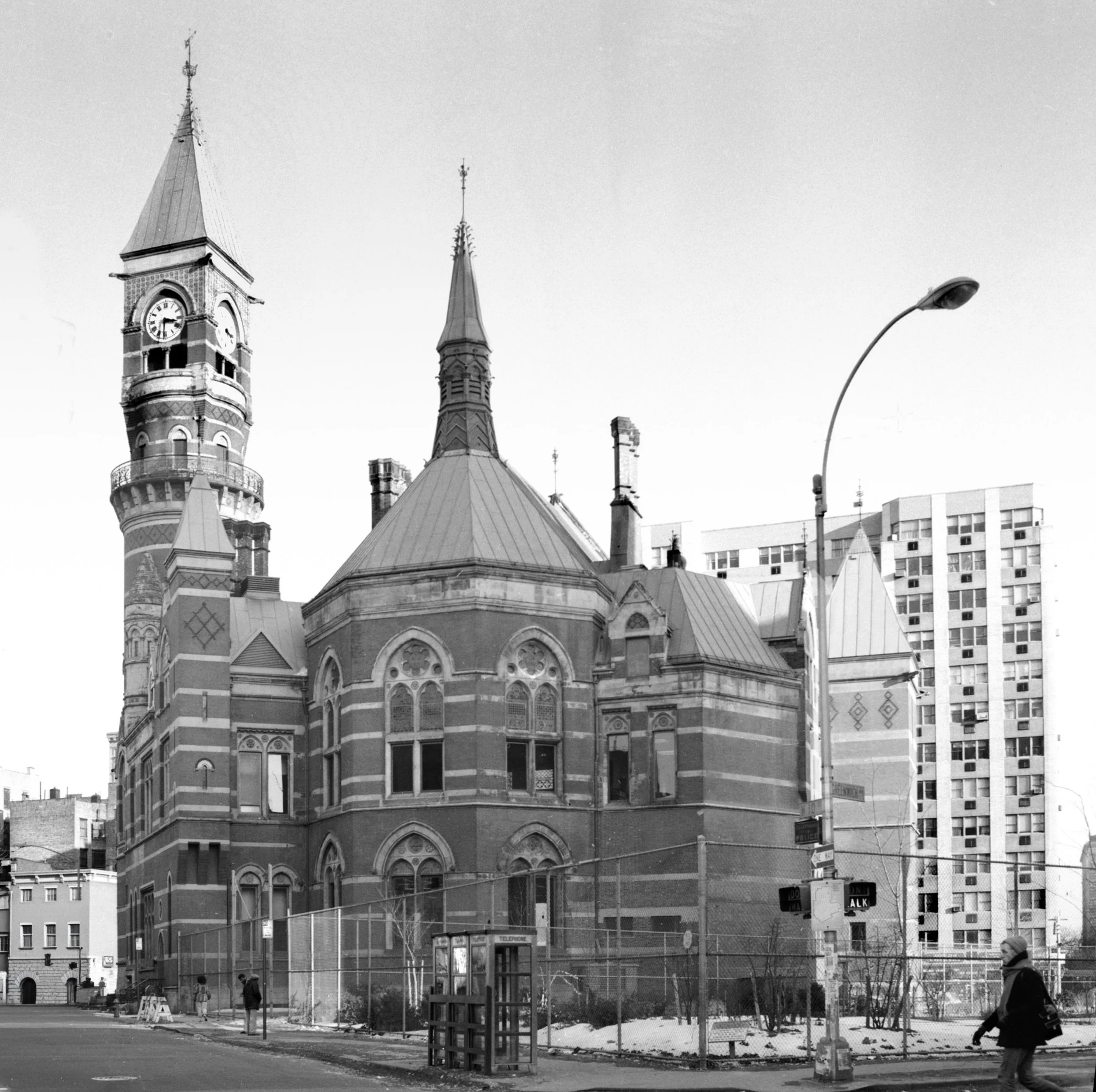 Black and white photograph of a historic church with two tall steeples, located at a street corner in an urban area, with a high-rise building and people walking in the foreground.