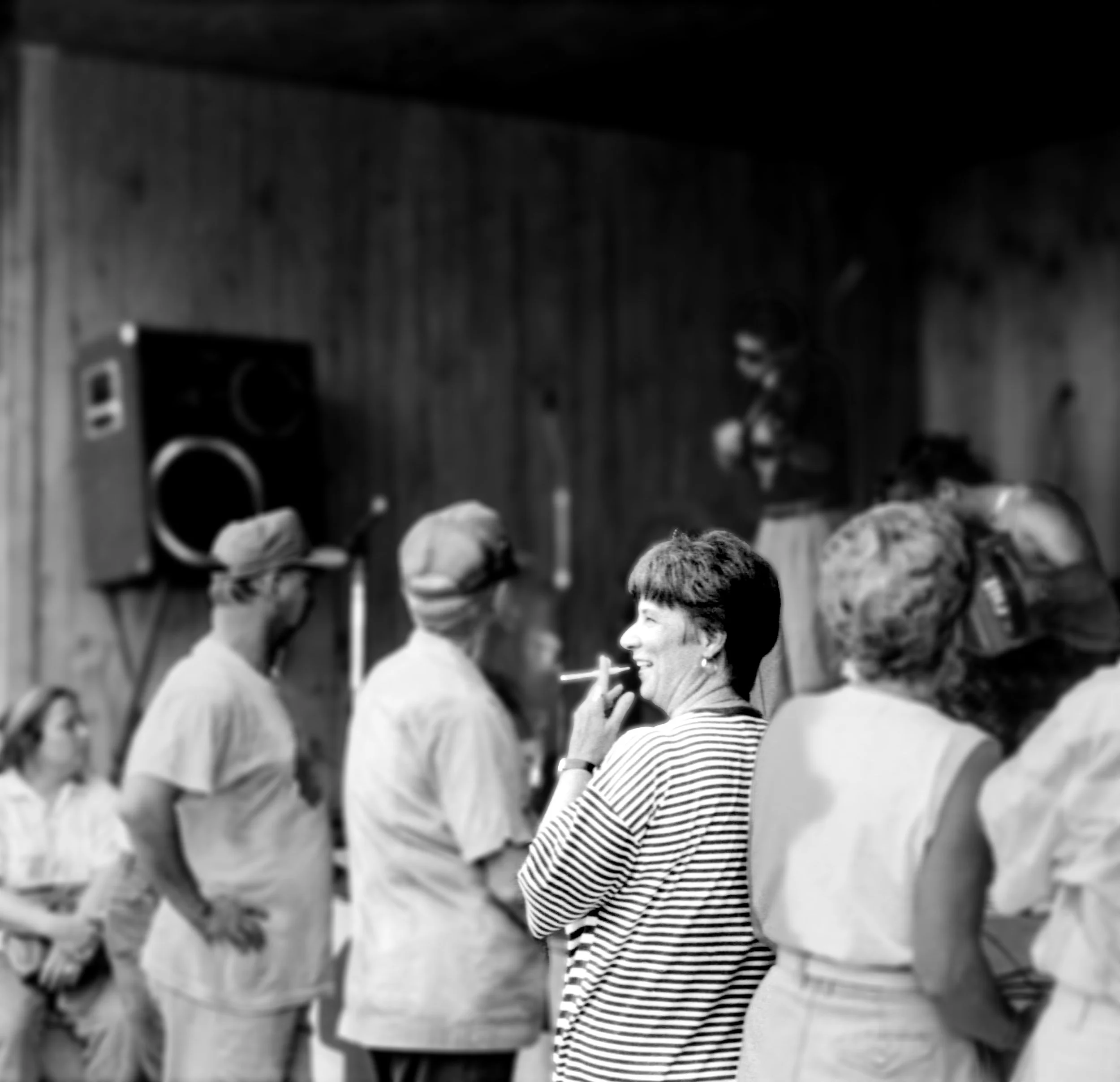 A group of people, including a woman smoking a cigarette, standing and talking in a room with wooden walls and a large speaker in the background.