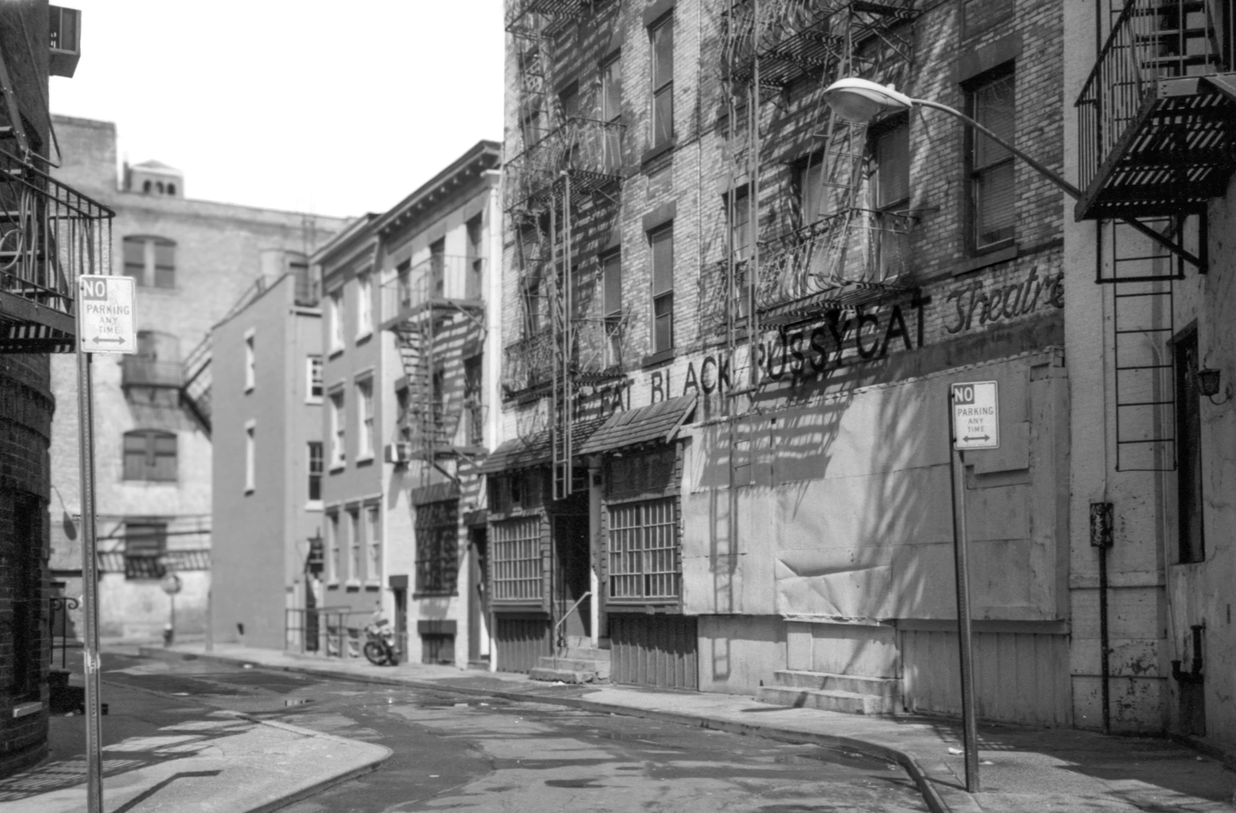 Black and white photo of an urban alley with fire escapes on brick buildings, street lamps, and a 