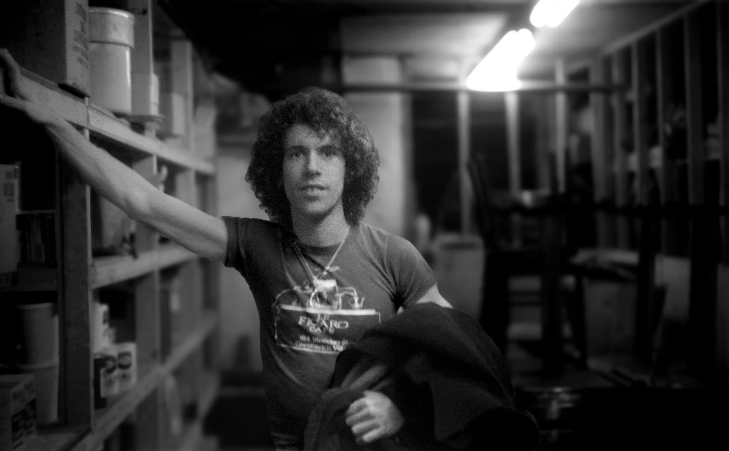 A person with curly hair wearing a t-shirt, standing in a storage or warehouse area with shelves and chairs, holding a jacket in one hand, and reaching with the other hand.