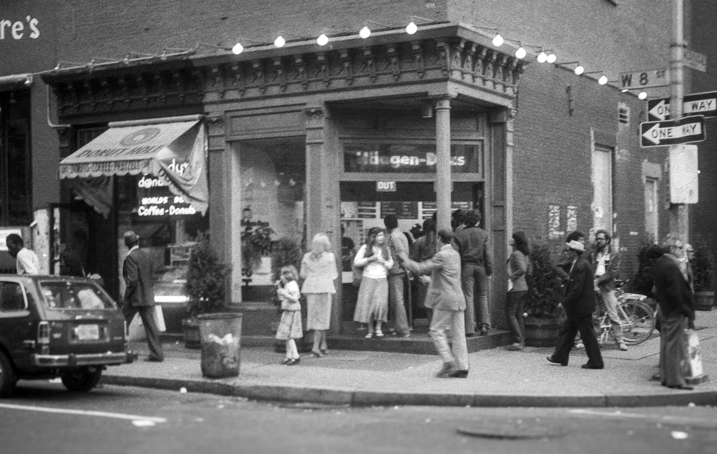 Black and white photo of a busy city street with people walking and interacting outside a shop. There is a car parked in the foreground and street signs indicating one way and a cross street.