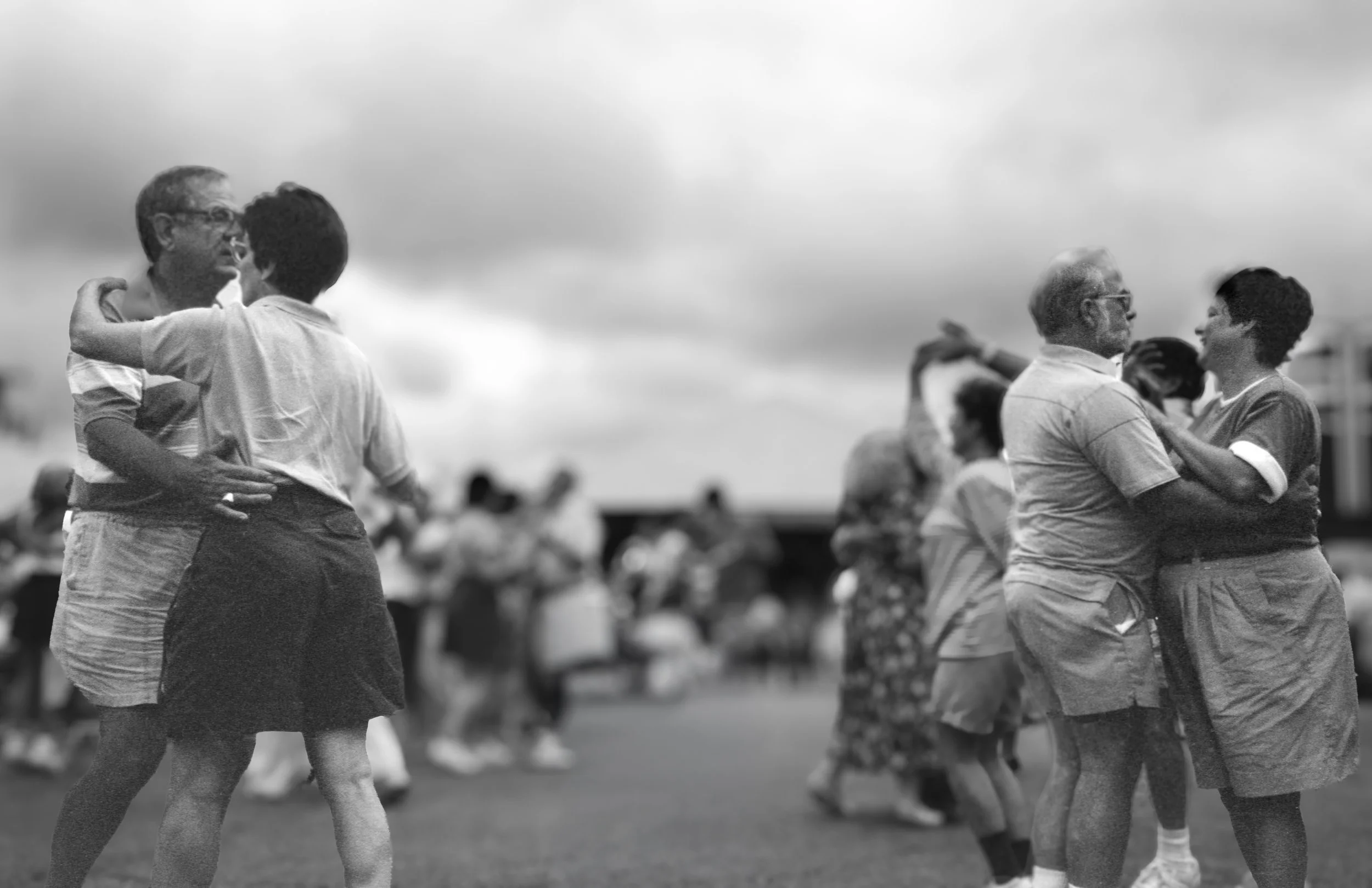 Couples dancing outdoors in pairs, with other people dancing in the background under an overcast sky.