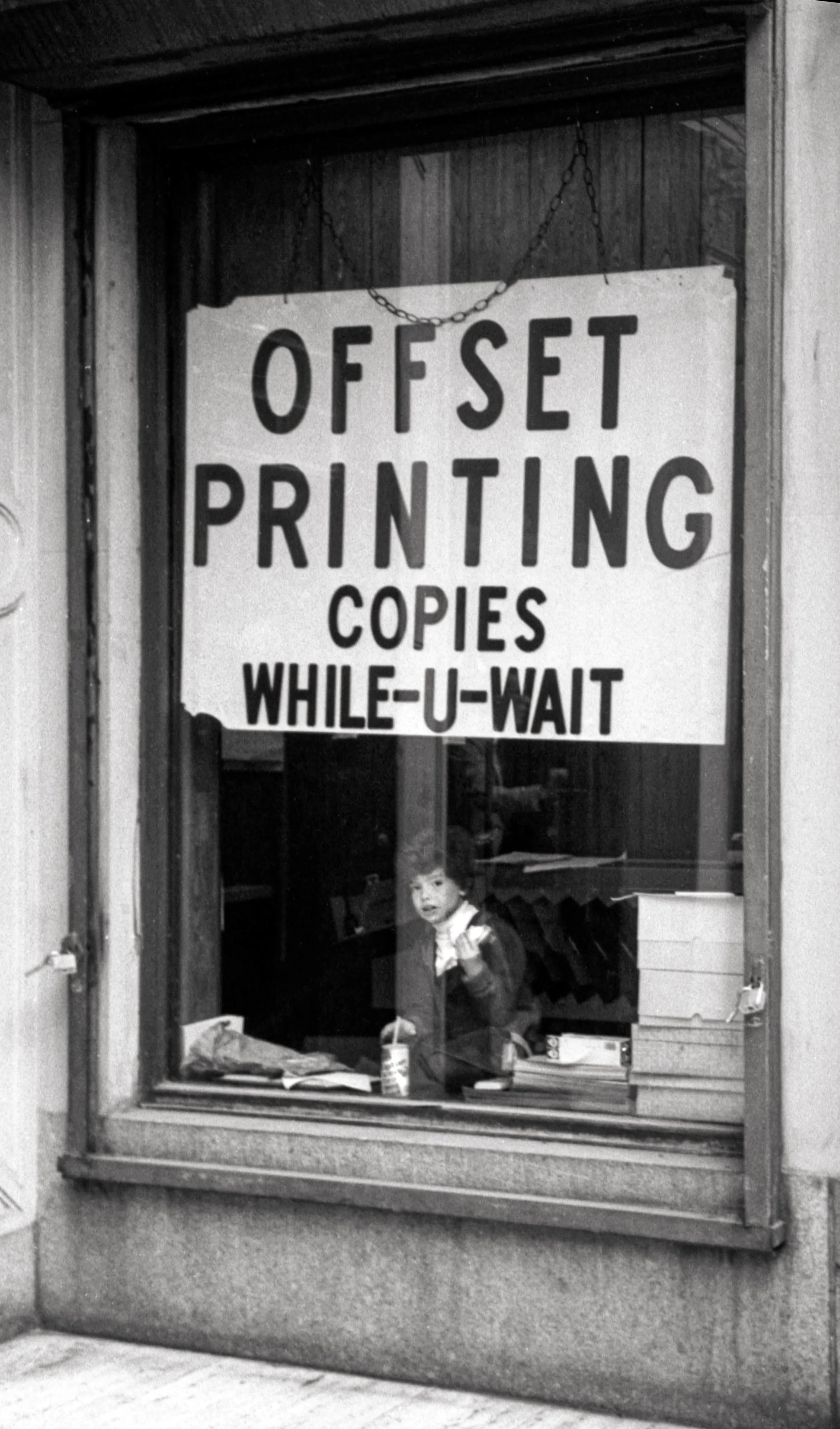 A black and white photo of a boy looking out a store window with a sign that reads "Offset Printing Copies While-U-Wait". The boy appears to be pointing or gesturing.